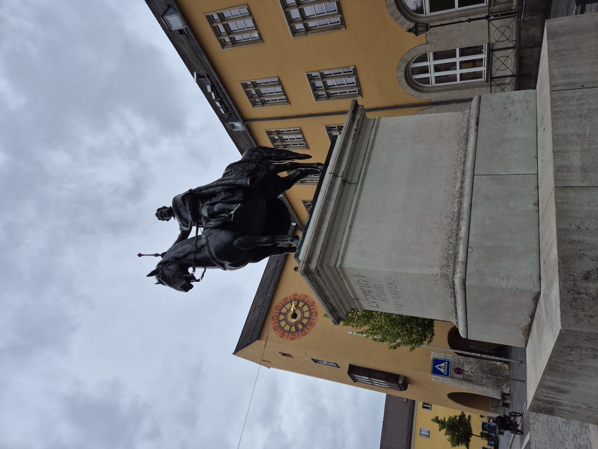 The image shows an equestrian statue of Ludwig I, King of Bavaria, in Regensburg, Germany. The statue is made of dark metal and stands on a stone pedestal. The pedestal is inscribed with "LUDWIG I KÖNIG VON BAYERN". In the background, there is a yellow building with a distinctive clock face. The clock has Roman numerals and astrological symbols. To the left of the statue, there is a pedestrian crossing sign and a red traffic sign. Further in the background, there are more buildings and trees. The sky is overcast. A person is visible in the lower left corner of the image, riding a three-wheeled bicycle. The scene appears to be a public square or street in a German city.