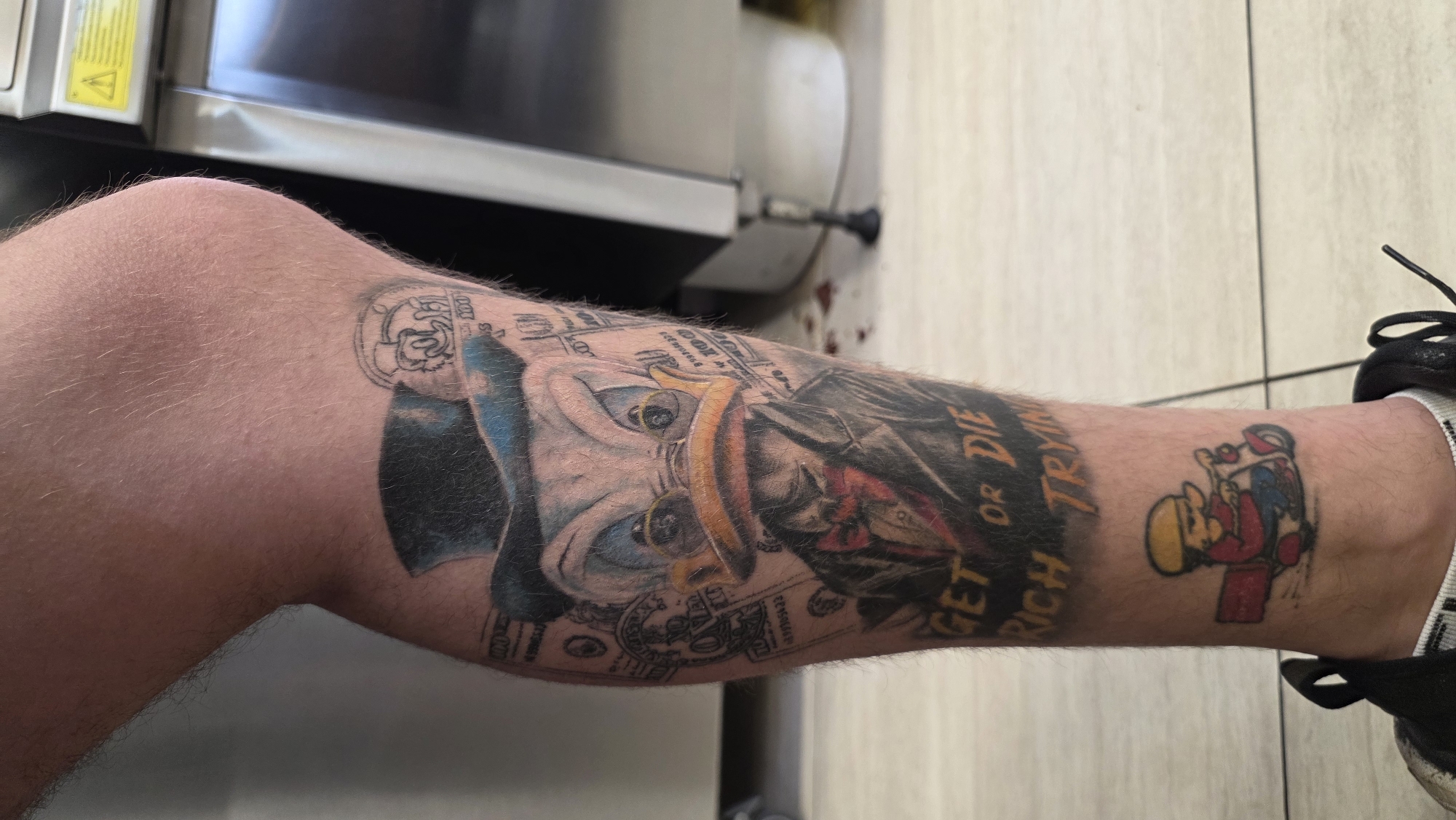 A lower leg is prominently displayed, featuring multiple tattoos. The primary tattoo appears to depict a face with dollar bills incorporated into the design, alongside the phrase "GET RICH OR DIE TRYIN." Another tattoo near the ankle shows a cartoon character. The leg is situated indoors, with a tiled floor and a stainless steel surface visible in the background. The person is wearing black and white athletic shoes. There is no location or detail to support that this image was taken in the city of Burgas, Bulgaria.