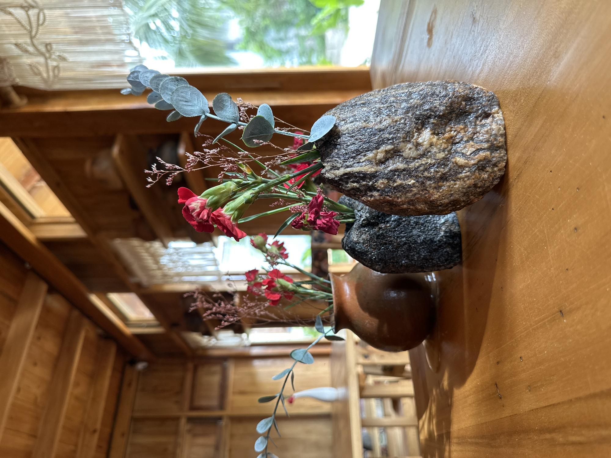 In a softly lit interior, likely a room in Phu Thuy Ward, Vietnam, a wooden table serves as a display for three floral arrangements. The foreground is dominated by the polished surface of the wooden table, reflecting the ambient light. Positioned on the table are three unconventional vases. The leftmost is a small, earthy brown ceramic pot holding a cluster of delicate red flowers and wispy dried branches. To its right, a rough, dark grey stone acts as a vase for a few more red flowers and a sprig of grey-green eucalyptus leaves. The largest vase, also a stone with varied grey and beige hues, is placed furthest to the right and holds a similar arrangement of red flowers and greenery. In the background, wooden walls and shelves create a rustic ambiance. A window with sheer white curtains allows diffused natural light to enter, offering a glimpse of lush green foliage outside, suggesting a tropical or subtropical environment. The overall scene is one of serene, naturalistic decoration.