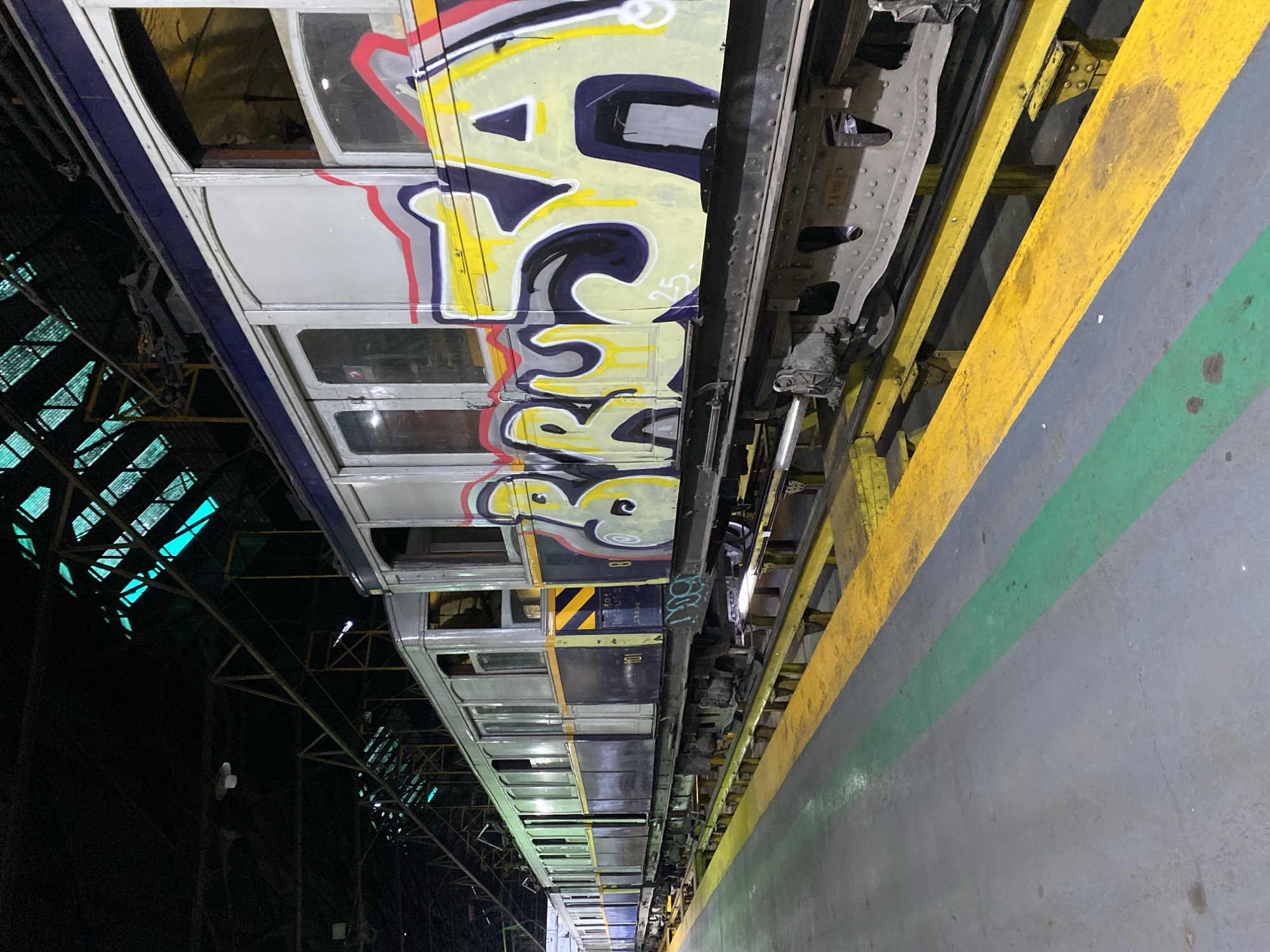An interior view within a maintenance depot in Buenos Aires, Argentina, displays a static line of historic subway cars. The car in the foreground exhibits a multi-tone livery of faded white, dark blue, and yellow stripes, extensively covered by graffiti featuring the stylized yellow, black, and red letters "BRUSA" or "BRUJA". Multiple arched windows are visible along the train's length. The cars' undercarriages are elevated and supported by yellow metal framework above a grey concrete floor, which bears a distinct green painted stripe. The upper environment reveals a dark, high-pitched roof structure with metal trusses and translucent blue-green panels, indicative of skylights. Overhead industrial light fixtures are also present. No individuals, active maintenance, or interactions are visible within the scene.