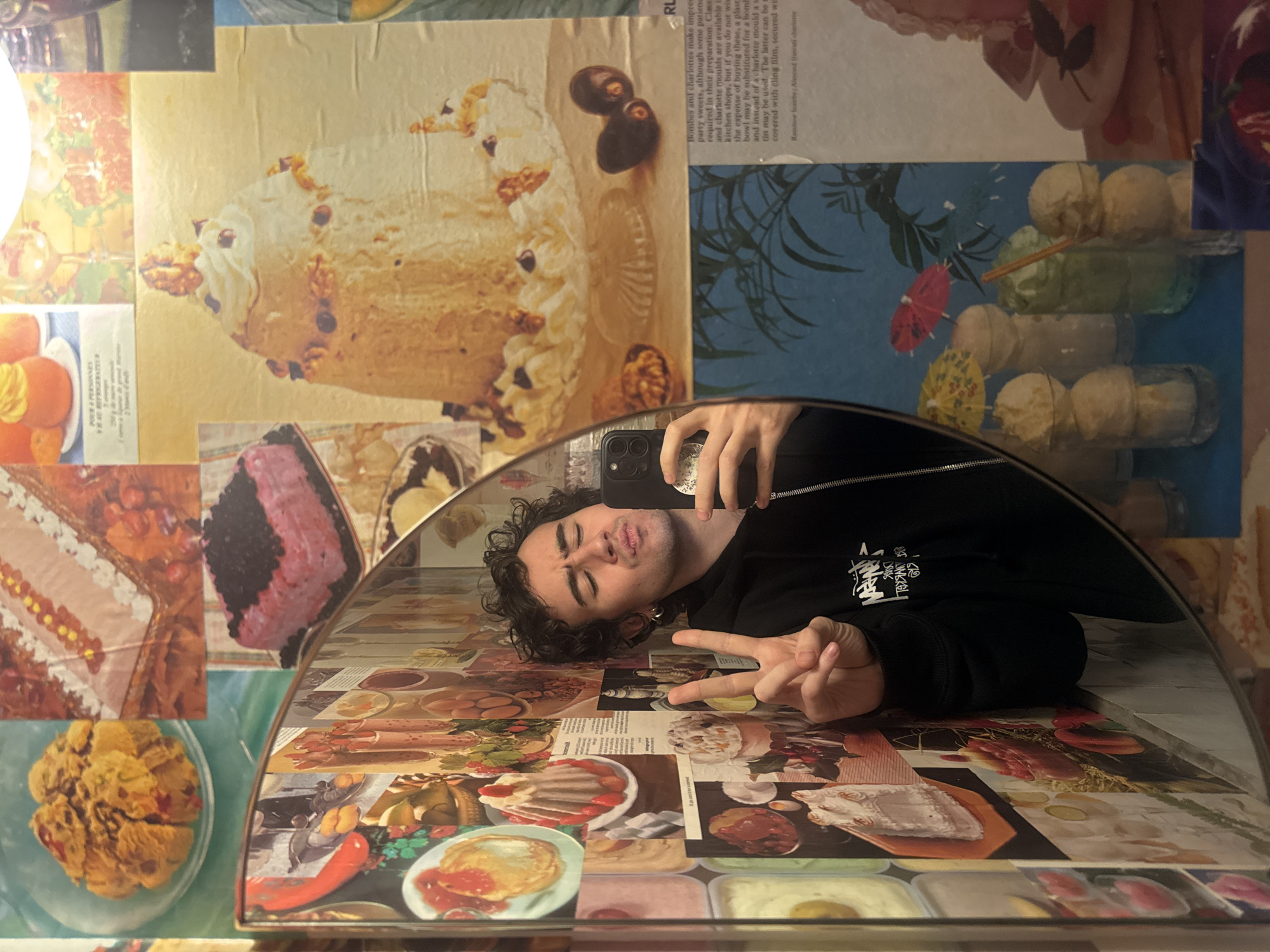 A person takes a mirror selfie. The person is a young man with curly brown hair, wearing a black hooded sweatshirt. He is making a peace sign with his right hand. The mirror reflects an image of him and a collage of food pictures in the background. The food pictures include cakes, desserts, cocktails, and other dishes. Text on the right side of the image is partially visible and written in English. There is no evidence that the image was taken in Paris.