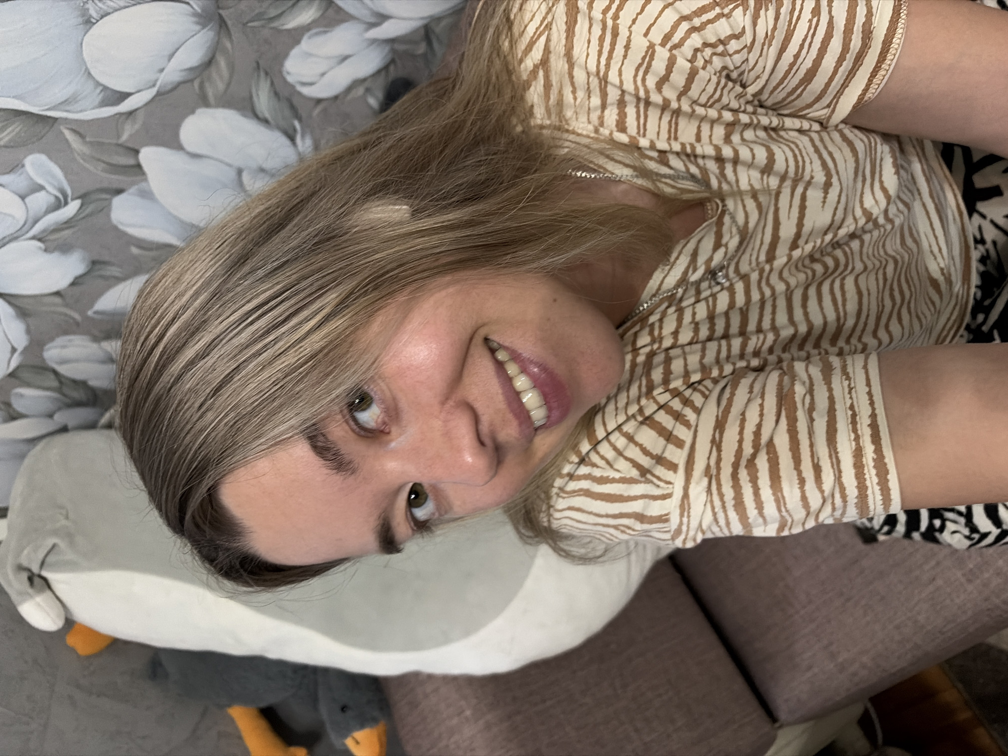 A light-skinned woman is depicted from the chest up, positioned indoors and looking directly at the viewer with a smile. She has light brown hair with darker roots, parted on her left side, and green eyes. Her eyebrows are dark and well-defined. She is wearing a cream-colored short-sleeved t-shirt with horizontal brown stripe patterns resembling animal print. A thin silver-colored chain necklace with a small pendant is visible around her neck. The woman appears to be seated on a light brown upholstered couch, a portion of which is visible to her left and at the bottom of the frame. Behind her head to the left is a large, light grey and white plush object, possibly a stuffed animal, with a visible orange beak at the top left corner, suggesting a stylized goose or duck. To her right, the background consists of grey wallpaper adorned with large white floral patterns. A small section of black and white patterned fabric is visible at the bottom of the image, beneath her right arm.