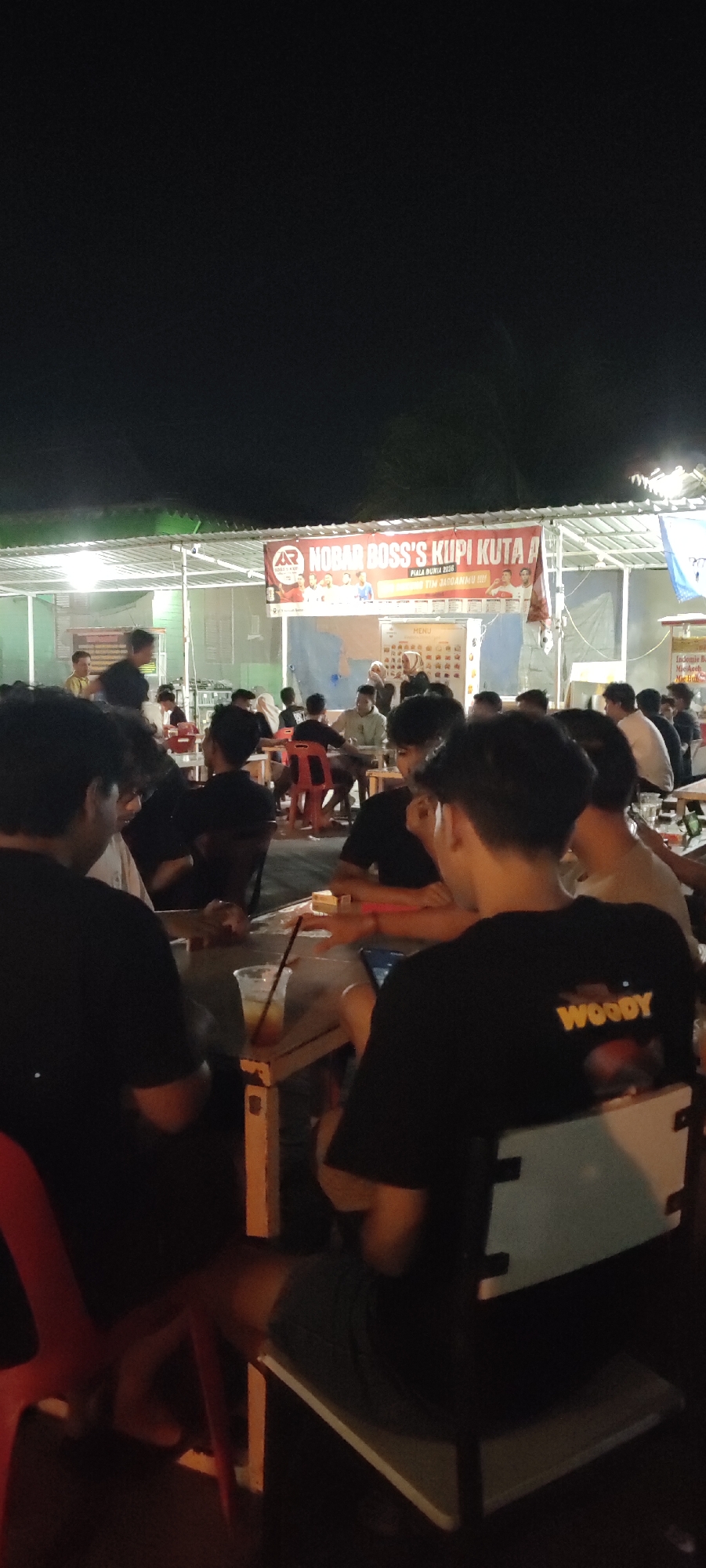 The image shows an outdoor gathering of people at night, likely at a cafe or warung in Banda Aceh, Indonesia. The scene is illuminated by artificial lighting from overhead structures and possibly string lights, creating a warm and inviting atmosphere. In the background, a banner with "NOBAR BOSS'S KUPI KUTA A PIALA DUNIA 2026 TIM JAGOANMU!!!" is prominently displayed, indicating a viewing event for the 2026 World Cup. A menu is also visible on a whiteboard.
The foreground is dominated by several individuals seated at tables, engaged in conversation and enjoying drinks. Most of the visible people are young men, dressed casually in t-shirts. One person in the foreground is wearing a black t-shirt with the word "WOODY" in yellow lettering on the back. The tables are made of wood and have plastic chairs. A cup with a drink and a straw is on one of the tables. The overall impression is that of a relaxed social gathering, possibly centered around watching a sporting event. The environment appears to be a semi-open-air establishment with a roof supported by poles.