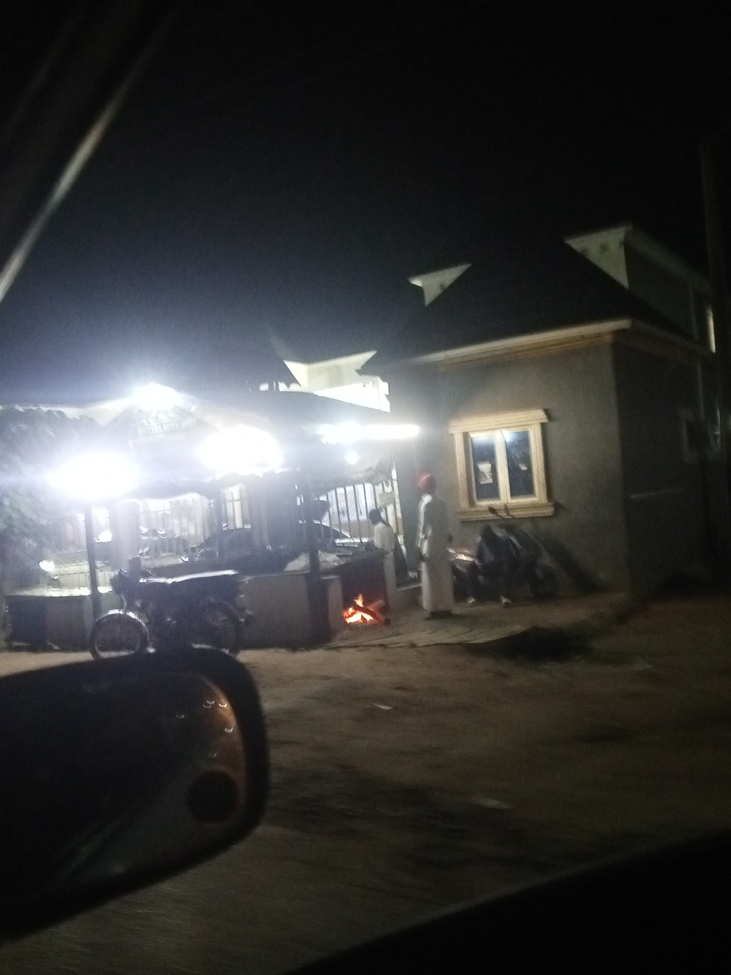 At night in the city of Unknown, Unknown, a fire burns near a building with a lit window. A motorcycle is parked nearby. Two people are standing close to the building. A structure with a light and a sign that reads "PERSATUAN SURIA" is also present. The image is taken from inside a vehicle, as indicated by the side mirror.