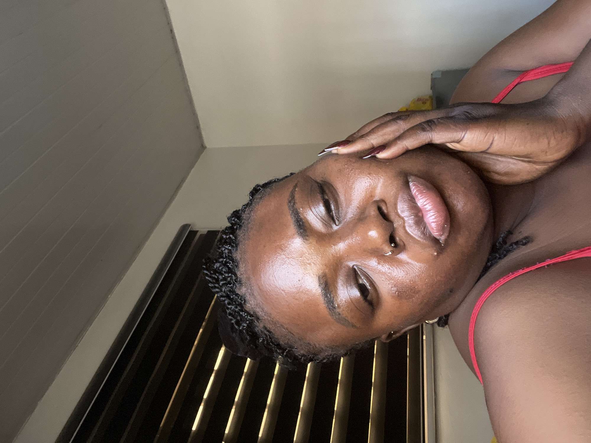The media file shows a close-up, tilted shot of a young Black woman. She is resting her forehead and temple on her hand, with her eyes closed and her lips slightly parted. She is wearing a red garment, likely a bikini top, visible on her shoulder and chest. Her hair is styled in small braids, and she has a nose ring and earrings. The background appears to be an indoor setting, with slatted blinds and a wall visible. The lighting suggests it might be daytime, with light coming from behind the blinds. The overall mood conveyed is one of relaxation or contemplation. There is no visible text in the image.