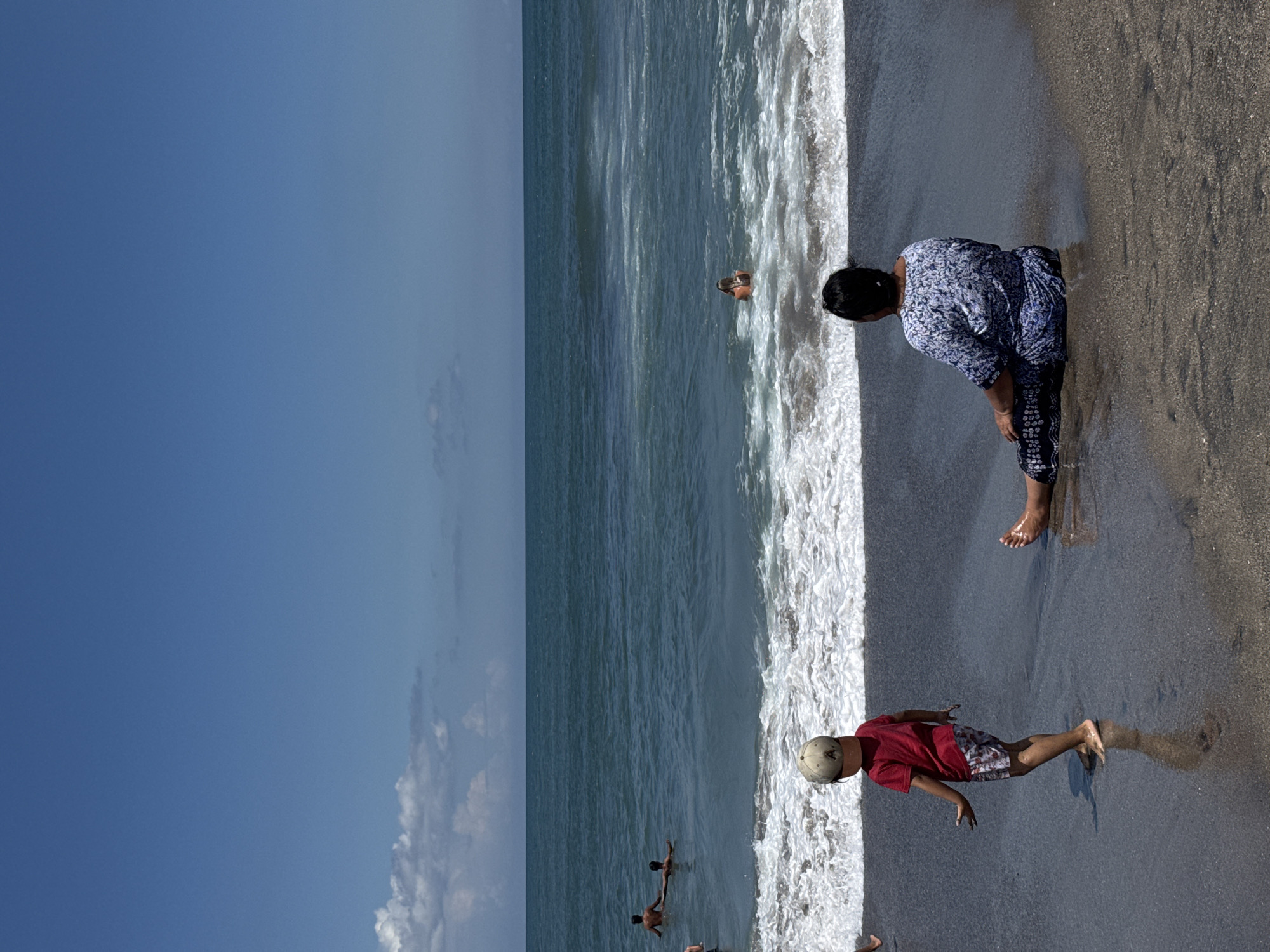 The image captures a scene on a beach. A woman in a patterned blue top and dark pants stands at the edge of the water. A young boy in a red shirt and patterned shorts plays near the water's edge. Other people are visible in the water. The sky is mostly clear and blue. The location is North Kuta, Indonesia.