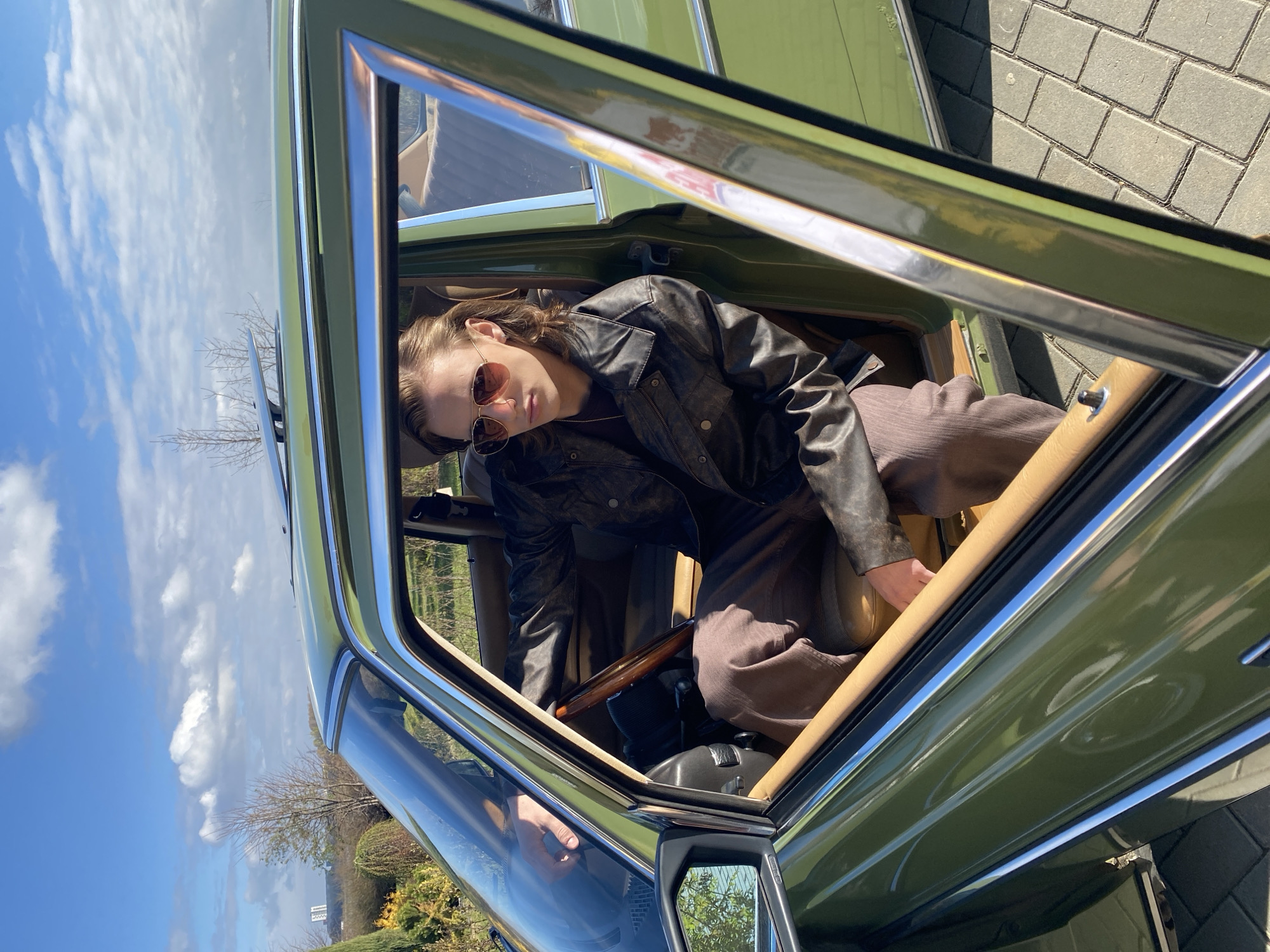 A person wearing sunglasses and a black leather jacket is reclining inside a green car with its door open. The person is wearing brown pants. The car's interior is tan. Outside the car, there is a view of a blue sky with scattered clouds, trees, and a paved area. The location cannot be determined as Yekaterinburg, Russia from this image alone.