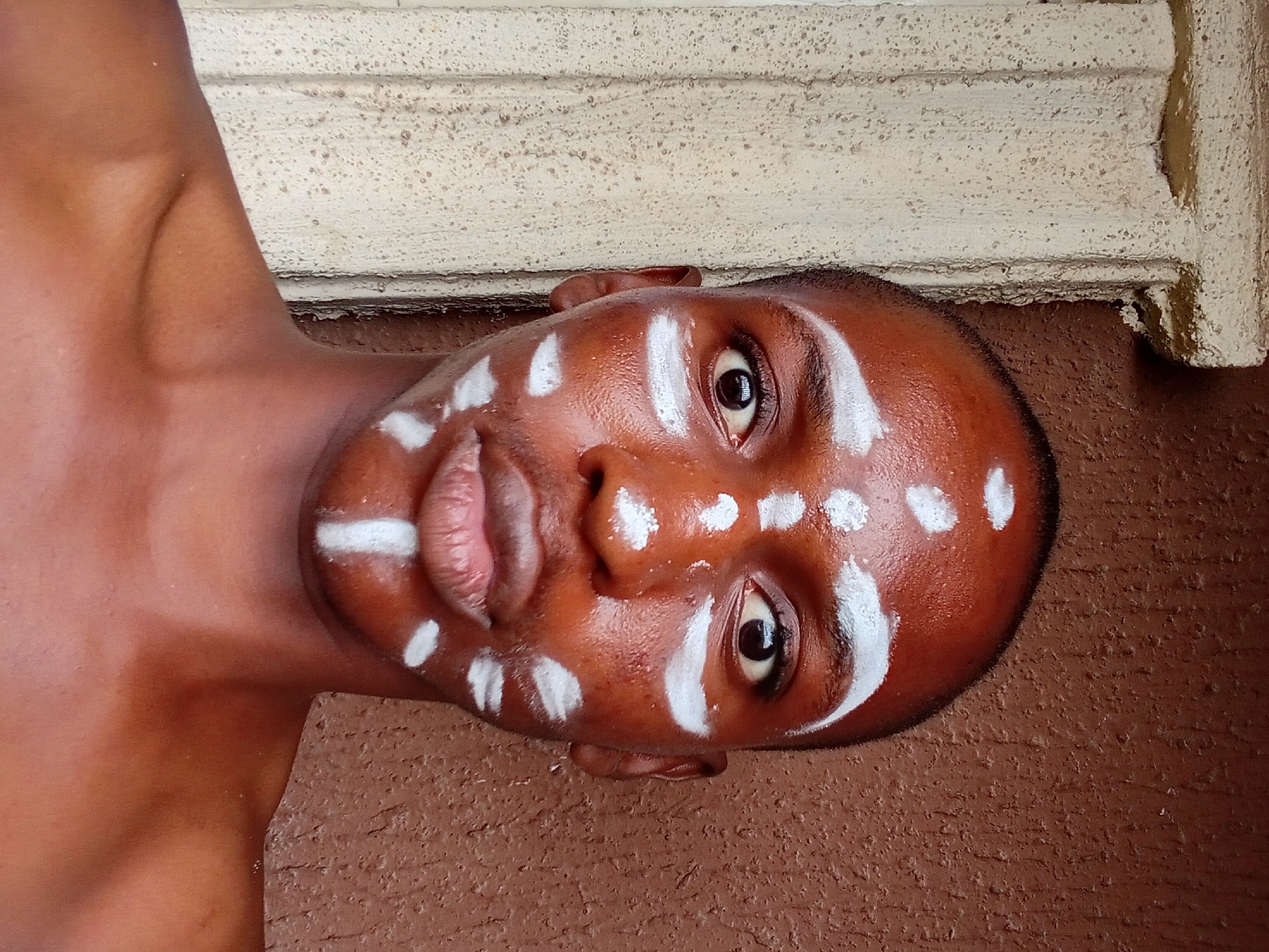 A close-up, frontal portrait captures a young Black individual adorned with white face paint, posing outdoors. The individual's head is shaved, and white paint is applied in circular patterns on the forehead and cheeks, with a vertical stripe on the chin and a horizontal stripe across the lower lip. The individual's skin is a warm, dark brown, and appears slightly dewy. Their eyes are open and directed towards the camera, and their lips are slightly parted.
The background is divided into two distinct textures and colors. To the left, a pale, textured surface, possibly a wall or column, features rough, speckled detailing. To the right, a reddish-brown, textured wall, likely a stucco or concrete finish, fills the remainder of the background. The lighting suggests it is daytime, with a soft, diffused light illuminating the subject's face. The overall impression is that of a cultural or artistic expression, possibly for a photoshoot or ceremony.
While the image itself does not contain explicit visual cues for its specific location within Nkpor, Nigeria, such artistic face painting traditions are common in various Nigerian cultures. The textured walls are also consistent with typical architectural elements found in residential or public spaces within Nigerian cities.