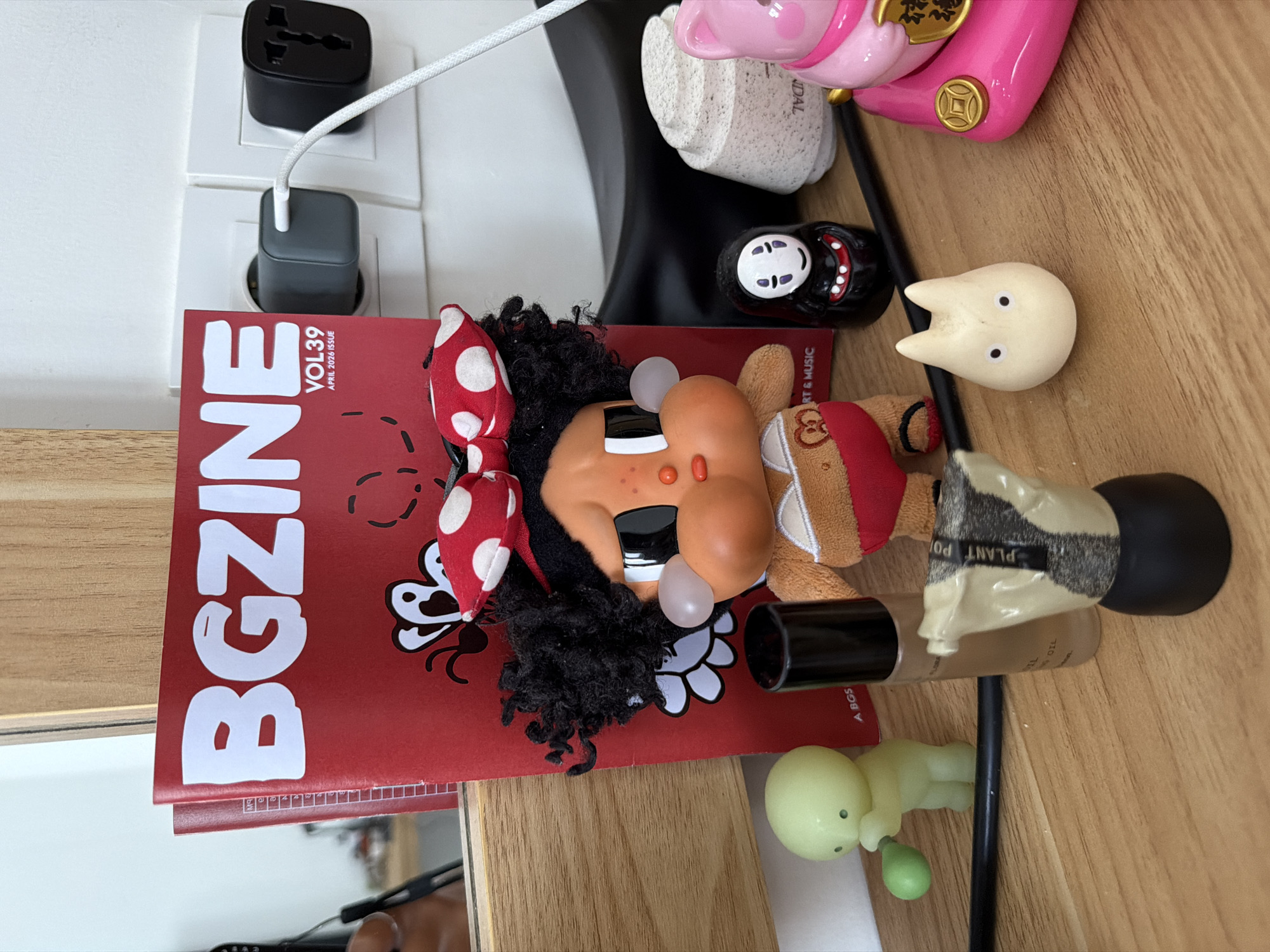 The image shows a collection of objects on a wooden surface. These include a red magazine titled "BGZINE VOL.39 APRIL 2023 ISSUE," a doll with a red and white polka-dot bow, and a black curly hairstyle, a black and white No-Face figure, a cream-colored cat figure, a green baby toy, and several beauty products including a tube with "PLANT POT" printed on it and a black-capped item. Two wall adapters are visible, one black and one gray, plugged into a white wall socket. The setting lacks specific geographical information that would allow identification of a location in Kuta Selatan, Indonesia.