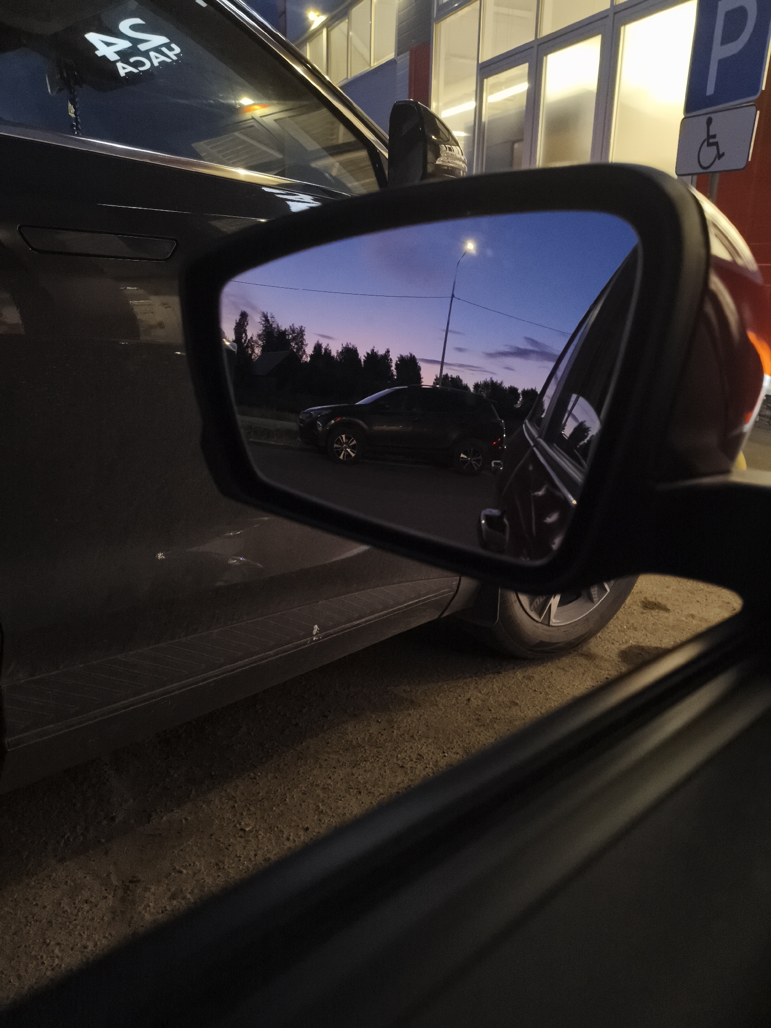 The image shows a view from inside a car looking out the driver's side window and into the side mirror. The time of day appears to be dusk, with a gradient of purple and blue in the sky.
In the side mirror, a black SUV is reflected. The SUV is parked on a paved surface, possibly a road or parking lot, with trees and a small building visible in the background. A street light is also reflected in the mirror, illuminating the scene.
Outside the car, the vehicle's dark gray paint is visible. On the rear window of the car, a sticker with white lettering that reads "AS ASAP" is applied. To the right of the car, a blue parking sign with a white wheelchair symbol is mounted on a pole, indicating a disabled parking spot. The building behind the sign has large windows with interior lights on.
The foreground shows the interior of the car, with the door frame and the edge of the car's interior panel visible. The ground beneath the car is rough and appears to be gravel or dirt. The image seems to be taken in a city or town in Russia, potentially near a building or parking area.