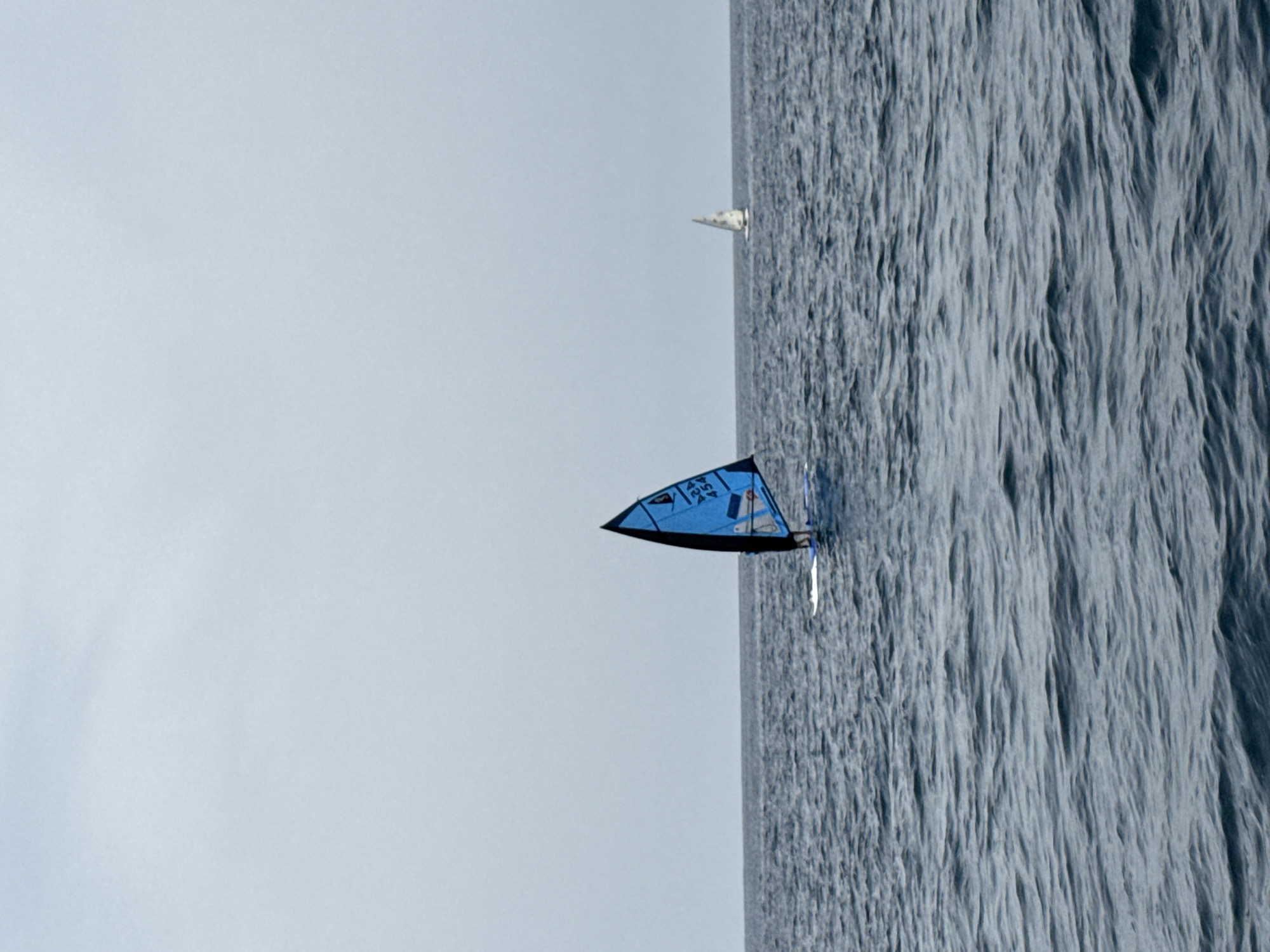 A windsurfer with a blue sail navigates the choppy, dark blue waters of the Mediterranean Sea, off the coast of Águilas, Spain. The sail features a black border and white lettering and numbers. The horizon is barely visible under a pale, overcast sky, and a second, distant sailboat with a white sail is visible on the right side of the frame. The water is characterized by small, consistent waves.