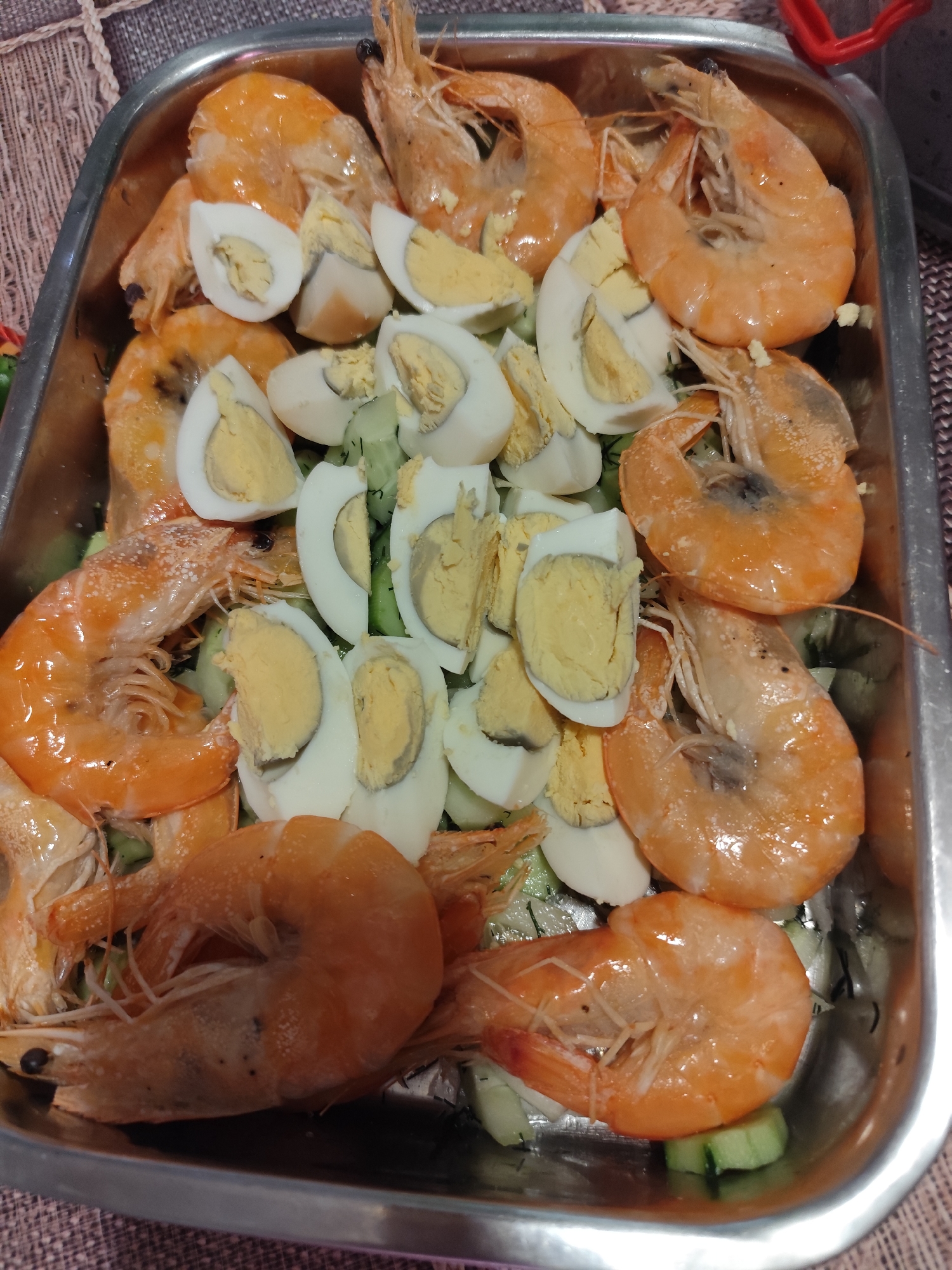 A close-up, top-down view shows a rectangular metal tray filled with cooked shrimp and sliced hard-boiled eggs. The shrimp are bright orange and appear to be whole, with their shells still on. They are arranged around the edges of the tray, with some overlapping. The hard-boiled eggs are cut into quarters and are scattered among the shrimp, with their yellow yolks visible. Thinly sliced green cucumber is visible underneath the shrimp and eggs, providing a bed for the ingredients. The tray rests on a patterned cloth, possibly a tablecloth or placemat, with a portion of a red object, perhaps a handle or a utensil, visible in the upper right corner. The scene is set indoors, and the lighting suggests it is daytime, though no specific geographical markers of Montana, Bulgaria are present in the image.