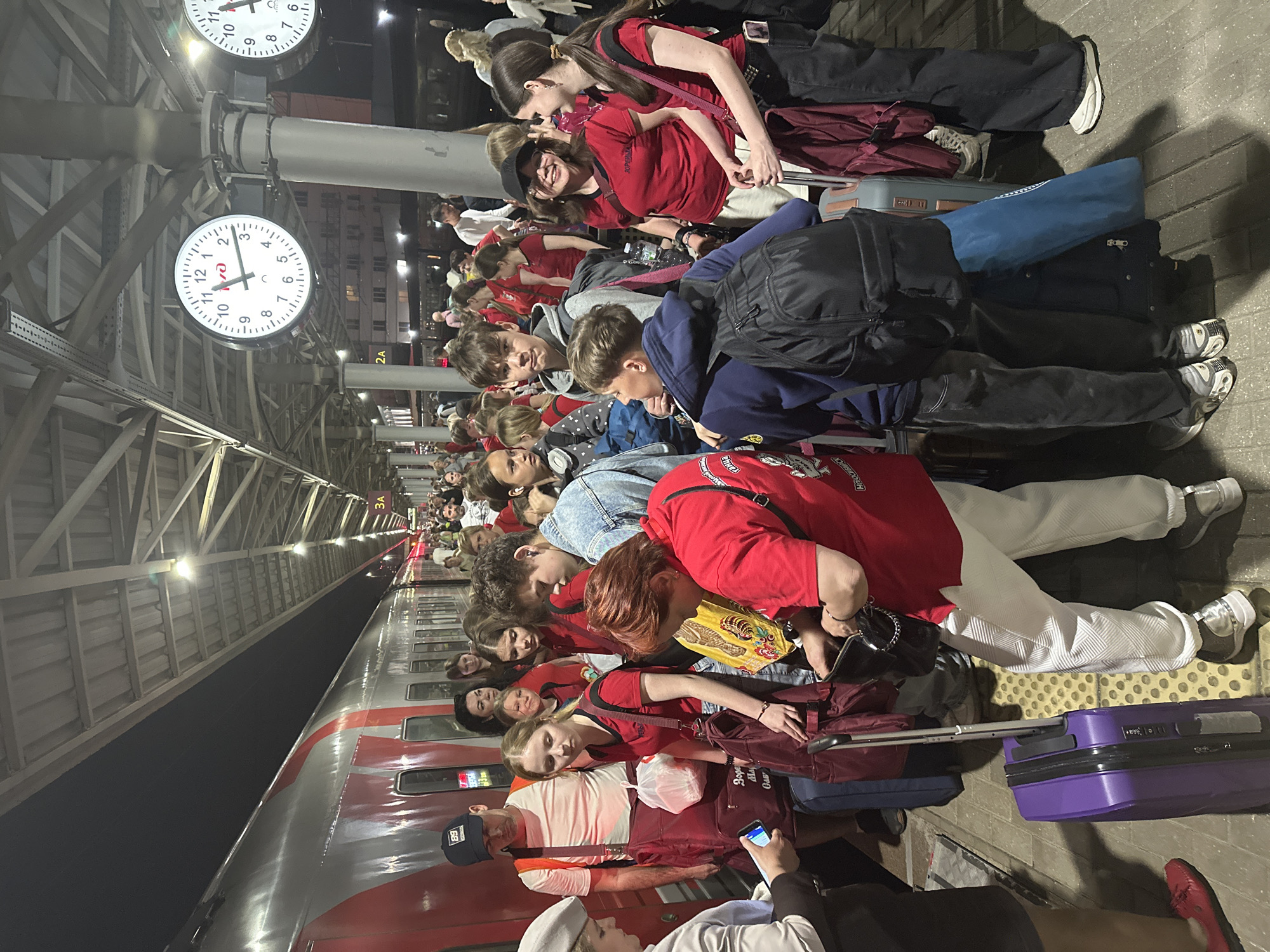 The image shows a crowd of people waiting at a train station, likely in Nizhny Novgorod, Russia. The train is visible to the left side of the image. Most of the people appear to be youths, many wearing red shirts and carrying luggage, suggesting they are traveling as a group. Some are seated on their luggage, while others stand or interact with each other. Two clocks are visible on the ceiling with time set around 1:10am.