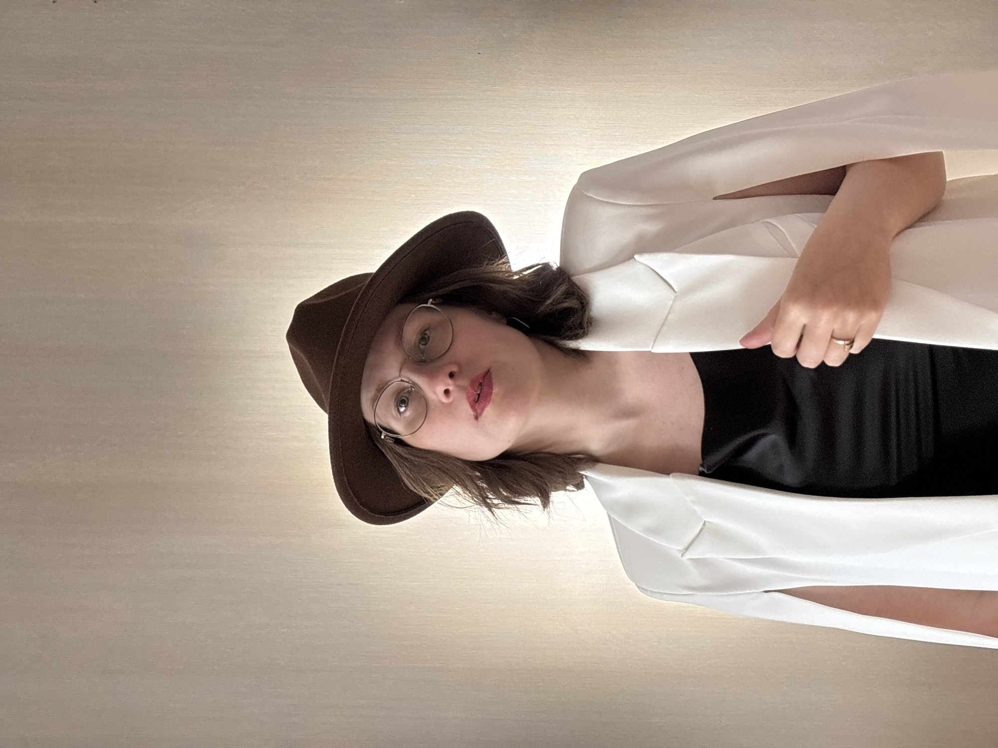 A woman wearing a brown hat and round eyeglasses is posing. She is dressed in a black top and a white jacket, and her left hand is visible, adorned with a ring. The location is unknown.