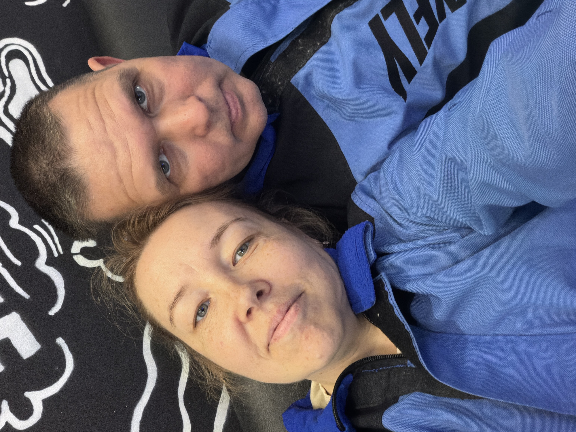 A selfie image shows two adults lying on a black surface with white patterns. The man at the top has short hair, fair skin, and is wearing a blue jacket with black lettering. The woman below has fair skin and is also wearing a blue jacket. The location is unknown and cannot be verified as Izhevsk, Russia, based on the visual information.