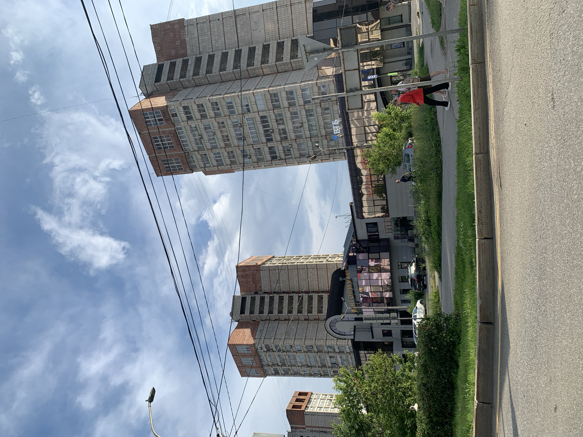 The image shows a street scene in Perm, Russia, featuring two tall, modern buildings with brick and concrete facades, and many windows. The buildings appear to be residential or mixed-use. A lower building with a glass facade connects the two taller structures. Several cars are parked on the street and in front of the buildings. People are walking on the sidewalk. Overhead, power lines crisscross the sky, and a lamppost is visible. The sky is partly cloudy with a bright sun. The foreground shows a paved road and a grassy area with bushes.