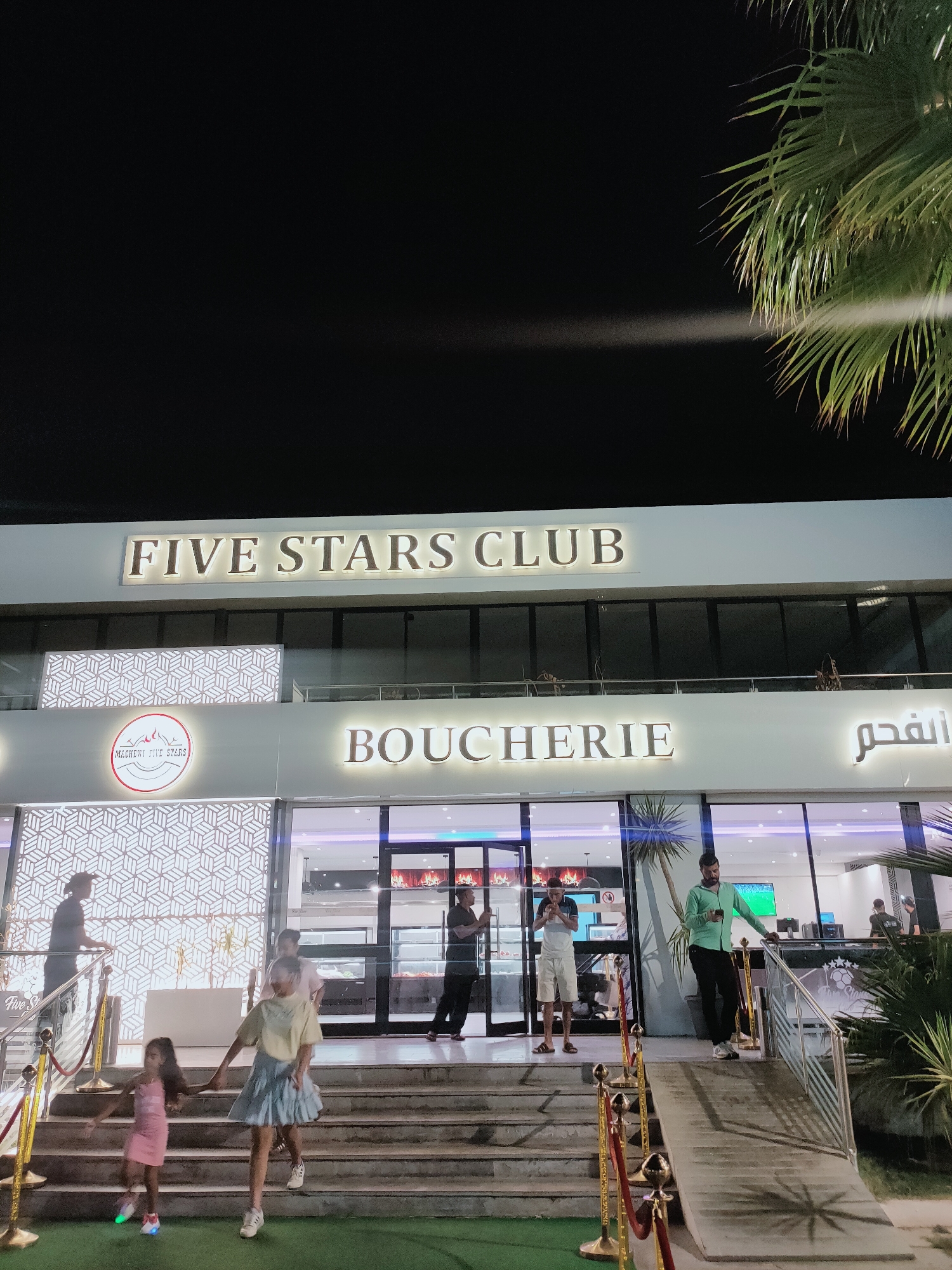 The image shows the exterior of a building at night, identified as "FIVE STARS CLUB" and "BOUCHERIE" (butcher shop) in Arabic. The building has a modern facade with illuminated signs. In front of the building, there are steps leading up to the entrance. Several people are visible, including a group of children walking down the steps in the foreground. A man in a green shirt is standing by a railing on a ramp. Other individuals are seen entering and exiting the establishment. The environment appears to be an outdoor public space, possibly a plaza or promenade, with palm trees and decorative barriers. The location is identified as Cebalat, Tunisia.