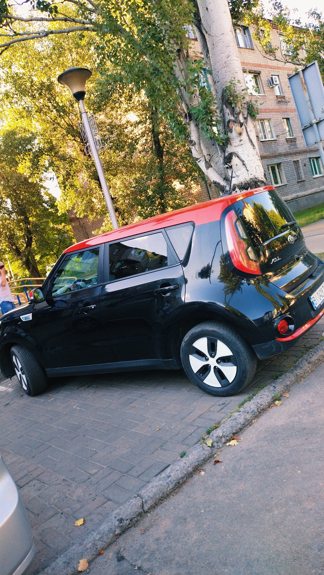 The image captures a black and red Kia Soul parked on a paved area next to a sidewalk in Horishni Plavni, Ukraine. The car is angled, with its driver's side facing the camera. The vehicle appears to be in good condition.
Behind the car, a building with brick construction and multiple windows is visible. A large tree with a pale, textured trunk grows prominently next to the building and over the car. The tree's branches, covered in green leaves, extend across the upper portion of the image. A streetlamp with a conical shade stands to the left of the tree. The ground in the foreground is a mix of paved walkway stones and asphalt, with a concrete curb separating them. Fallen leaves are scattered on the ground, suggesting an autumn setting.
In the background, a portion of another silver vehicle is visible on the far left of the image. To the left of the car, a person wearing a light-colored top and jeans is partially visible, standing near a yellow railing. The overall lighting suggests it is daytime, possibly late afternoon given the warm tones.