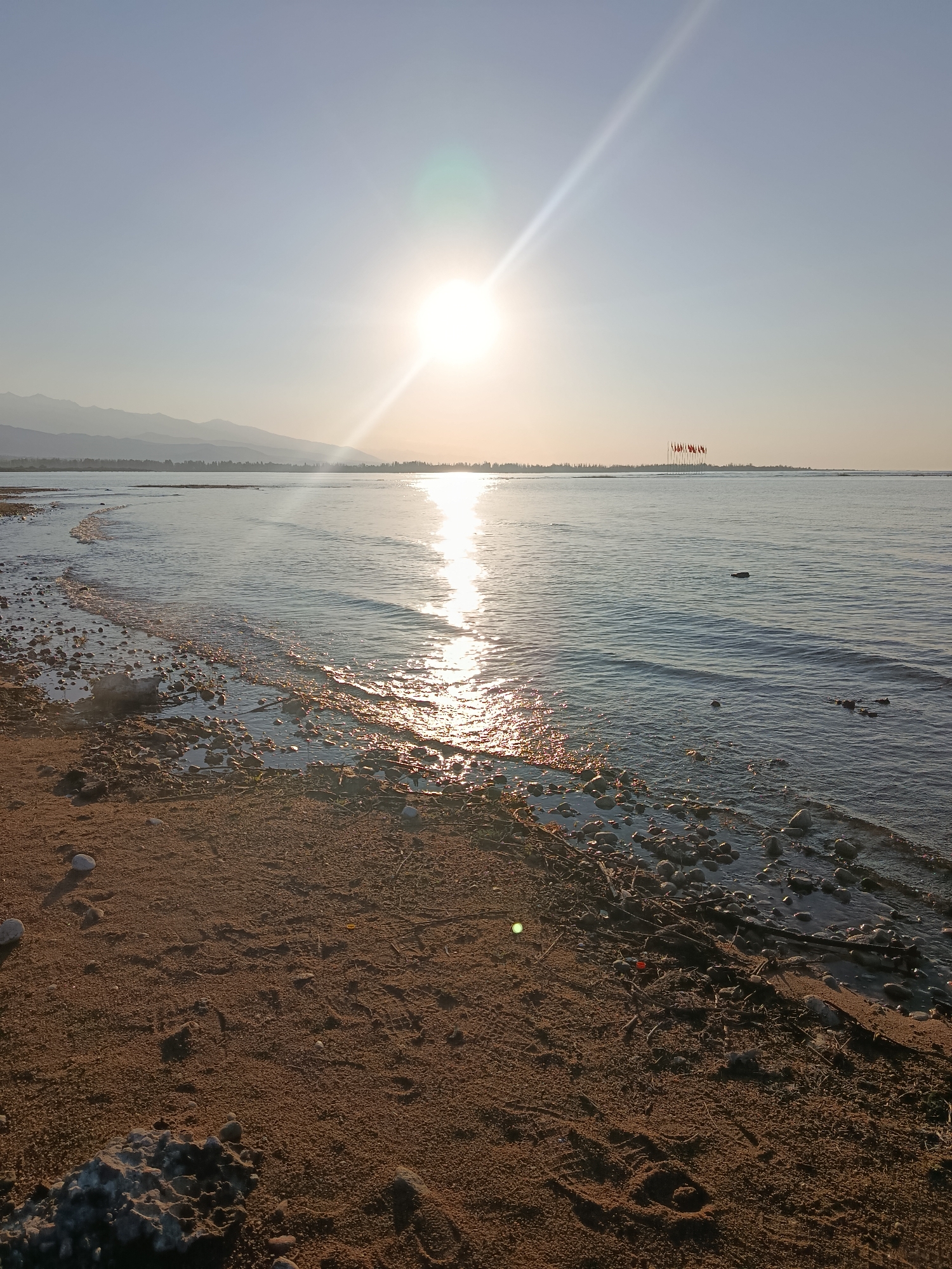 The image shows a tranquil lakeside scene in Kurskoe, Kyrgyzstan, with the sun setting over the water. The foreground consists of a sandy beach littered with rocks and driftwood. Gentle waves lap at the shore, reflecting the golden hues of the setting sun. In the distance, a line of mountains forms a silhouetted backdrop against the pale sky. A group of flags is visible on poles in the water. The overall atmosphere is peaceful and serene.