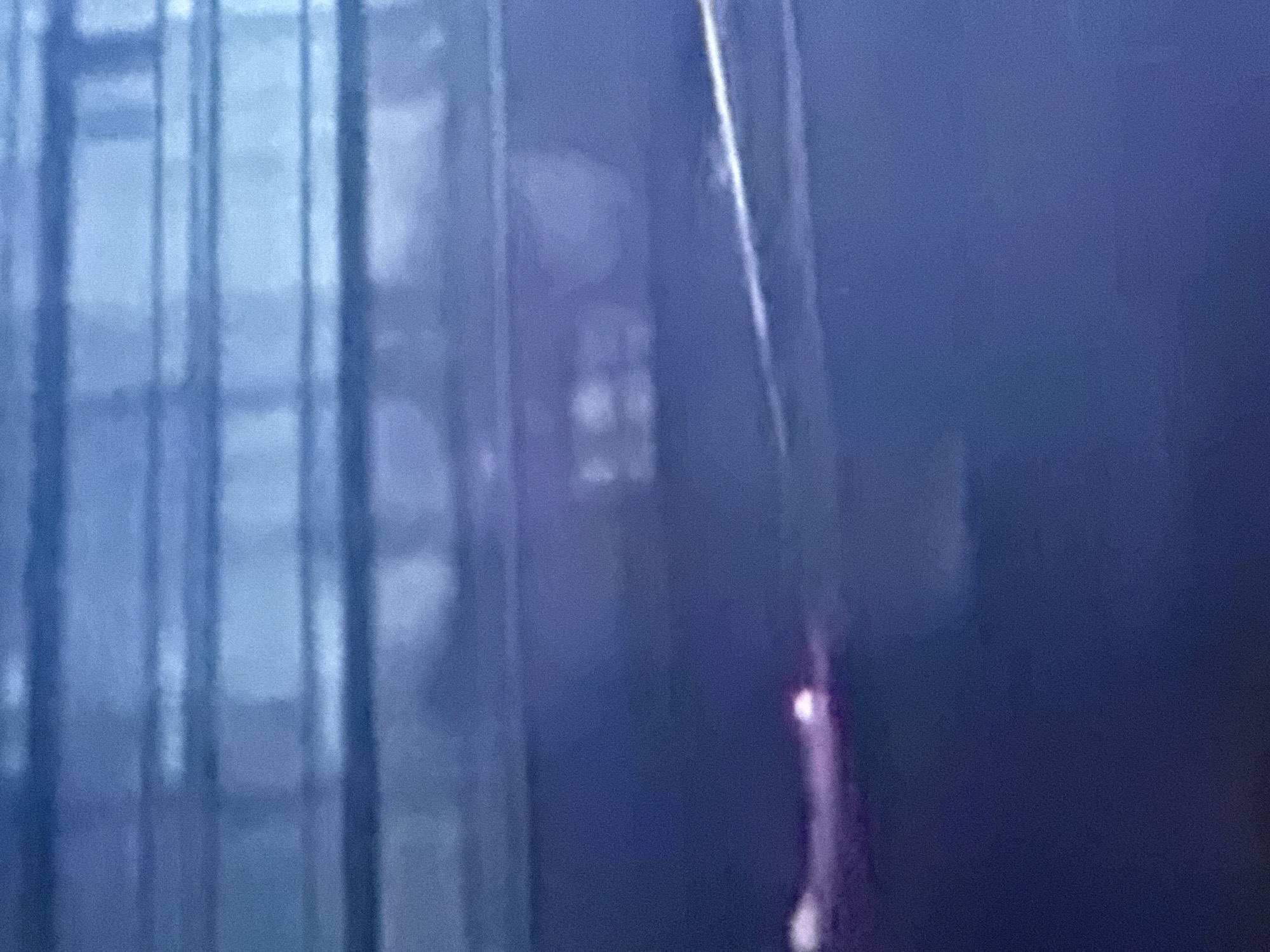 The image depicts a dark, blurry scene with horizontal lines suggestive of artificial lighting or reflections. In the lower left quadrant, a bright, pinkish-white light source is visible, possibly a reflection or a light bulb. Further to the right, a faint white line cuts horizontally across the frame, possibly indicating a surface or object. The overall impression is abstract and lacks clear subject matter or context. The provided context of Nyurba, Russia, does not offer any specific details that can be visually confirmed from the image.