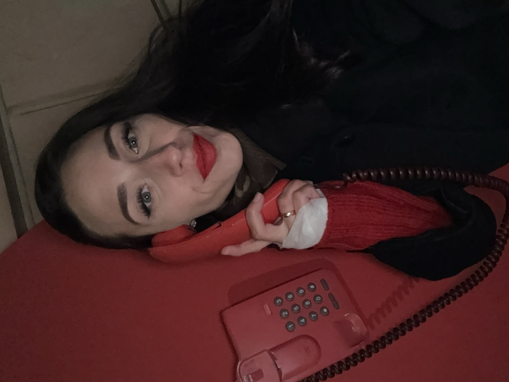 A woman with dark hair and red lipstick is lying down, holding a red telephone receiver to her ear. Her left hand is holding a white bandage. The telephone base is also red. The background is dark, and a portion of a wall is visible in the upper left corner. The location cannot be determined from the image.