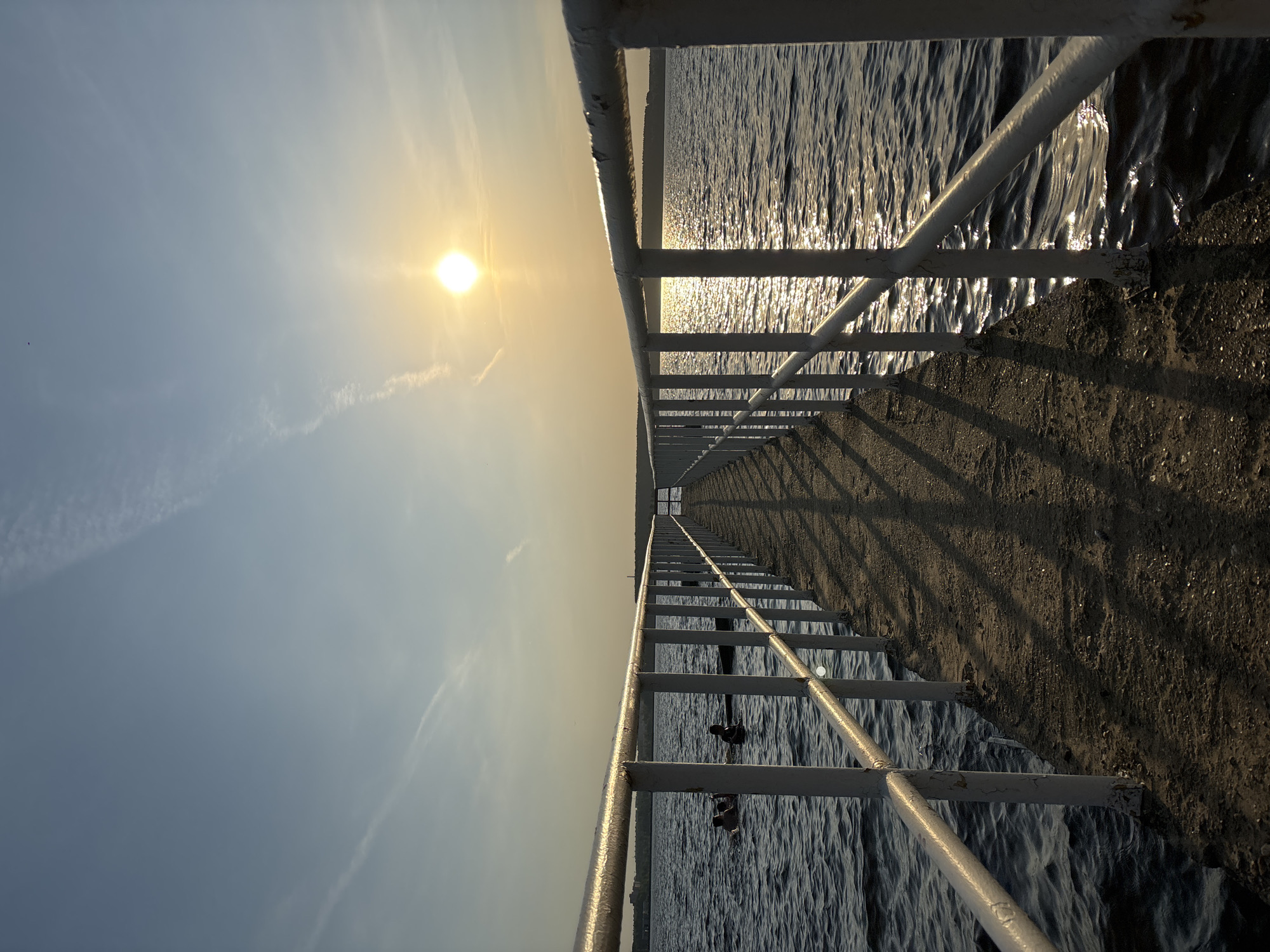 The image shows a long, narrow pier stretching into the water, framed by a white metal railing on either side. The sun is positioned directly ahead, creating a perspective that draws the eye toward the horizon. The water surrounding the pier has a rippled surface, reflecting the sunlight. The pier's concrete walkway is visible, with shadows cast by the railing. The sky is a gradient of blue and yellow where the sun is. There are some wisps of clouds in the sky. The pier is located in Perm, Russia.