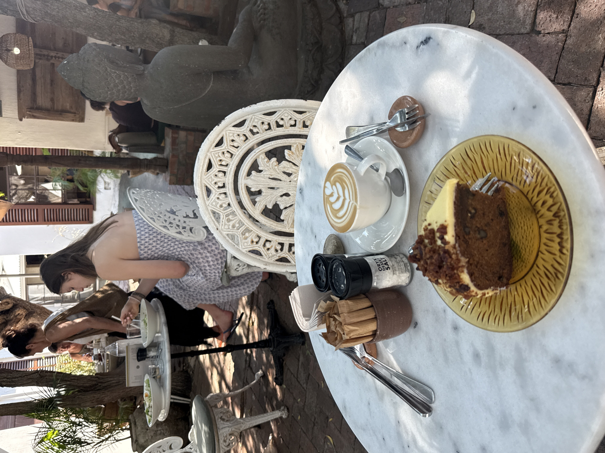 This image depicts a cafe scene in Cape Town, South Africa. A young woman with long brown hair is seated at a white, ornate wrought-iron chair and is eating a salad. The table is white marble. On the table is a plate with a slice of cake, a cup of coffee, salt and pepper shakers, and cutlery. Other people are seated at nearby tables, enjoying meals. In the background, there's a large statue of a Buddha and various plants and trees that provide shade. The architecture appears to be in a colonial style, indicative of certain areas in Cape Town.