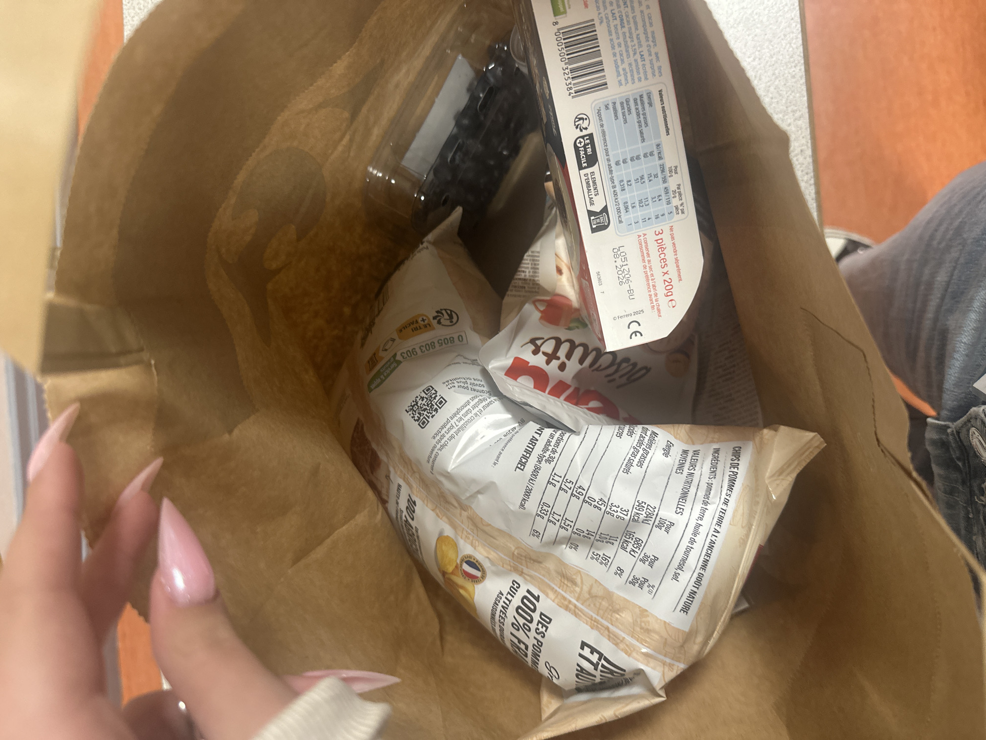 A brown paper bag is seen from above, filled with food items including potato chips, a box of Nutella biscuits, a plastic container of blueberries, and a package of what appears to be chocolate bars. A person’s hand with long pink fingernails is visible on the left side of the image. The background is a mix of colors including brown, orange, and gray, suggesting the image was taken indoors, likely on a table. Based on the text on the packaging, the location is likely Strasbourg, France, but this is not confirmed by the image.