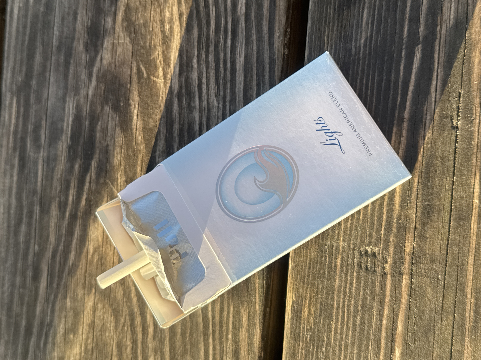 An open pack of light blue and white cigarettes rests on a weathered wooden surface, likely a table or bench. One white-filtered cigarette is partially extended from the top opening of the pack. The wooden surface displays prominent vertical grain patterns and varying shades of brown and grey, suggesting exposure to elements.
The scene is illuminated by strong, low-angle sunlight, casting distinct, elongated shadows across the wooden texture, indicative of either early morning or late afternoon. The light highlights the subtle gloss of the cigarette packaging and the rough grain of the wood. There are no people or other objects visible in the frame, focusing entirely on the cigarette pack.
Visible text on the pack includes "Lights" in an elegant cursive script, and "PREMIUM AMERICAN BLEND" in a smaller, uppercase font along the side. Inside the opened pack, on the silver foil lining, the word "Pull" is visible. A stylized logo resembling a wave or flame, rendered in blue and silver, adorns the center of the pack. The image captures a singular object in an outdoor setting, presumably in London, United Kingdom, under specific lighting conditions.