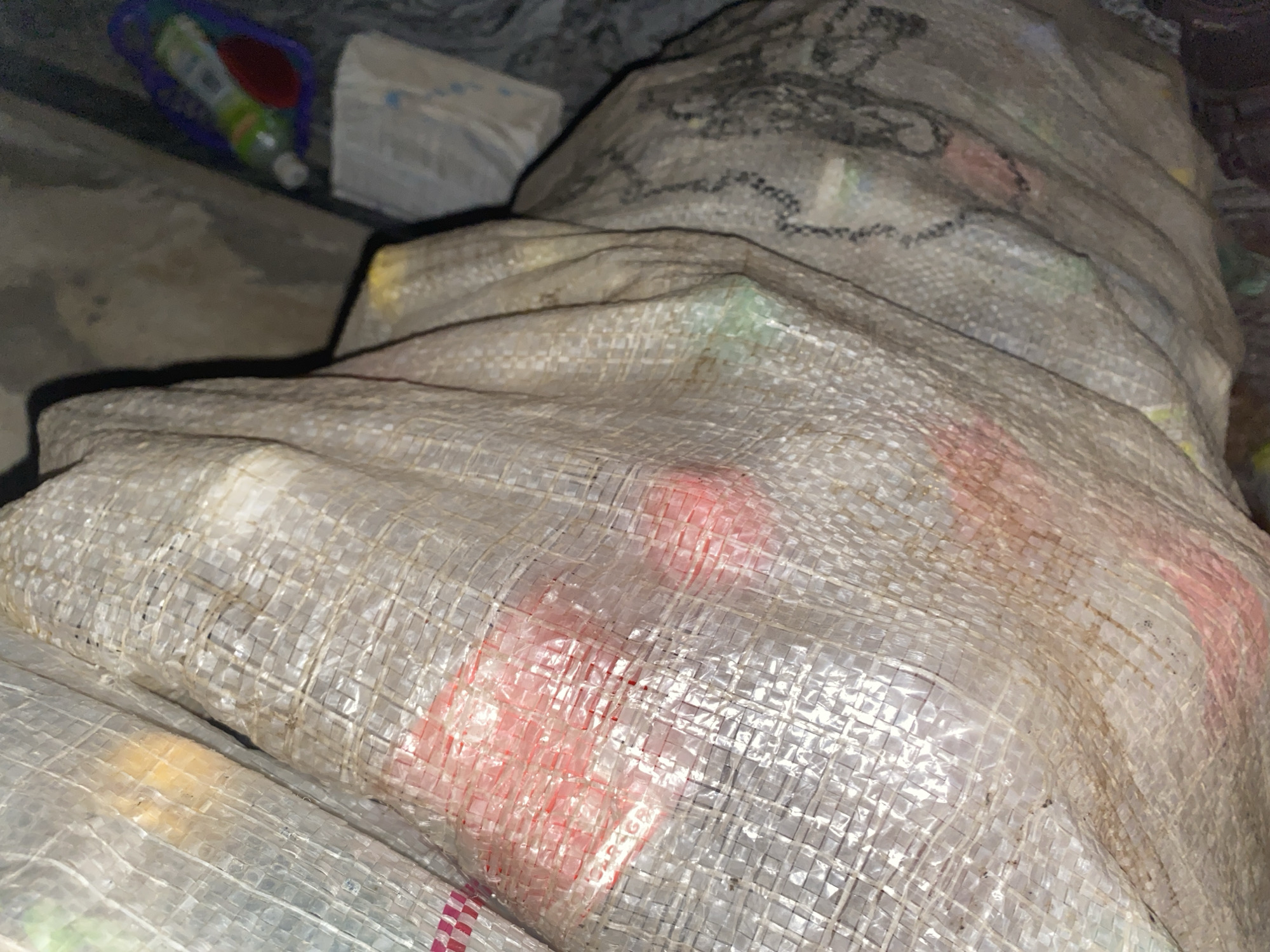 The image shows a close-up view of several large, opaque plastic sacks filled with what appears to be recyclable materials. The sacks are made of woven plastic and are somewhat translucent, allowing glimpses of the contents within. Some of the sacks have visible reddish or pinkish colored items, and some lighter yellow or orange items. The sacks are stacked together, and the lighting suggests an indoor, possibly storage or collection area. In the upper right corner of the image, partially obscured, are a few other items, including a plastic bottle with a colorful label and a white object that might be another bag or container. The overall environment appears to be somewhat cluttered and dimly lit. There are no people visible in the image. The location is not specified beyond the city of Unknown, Unknown.