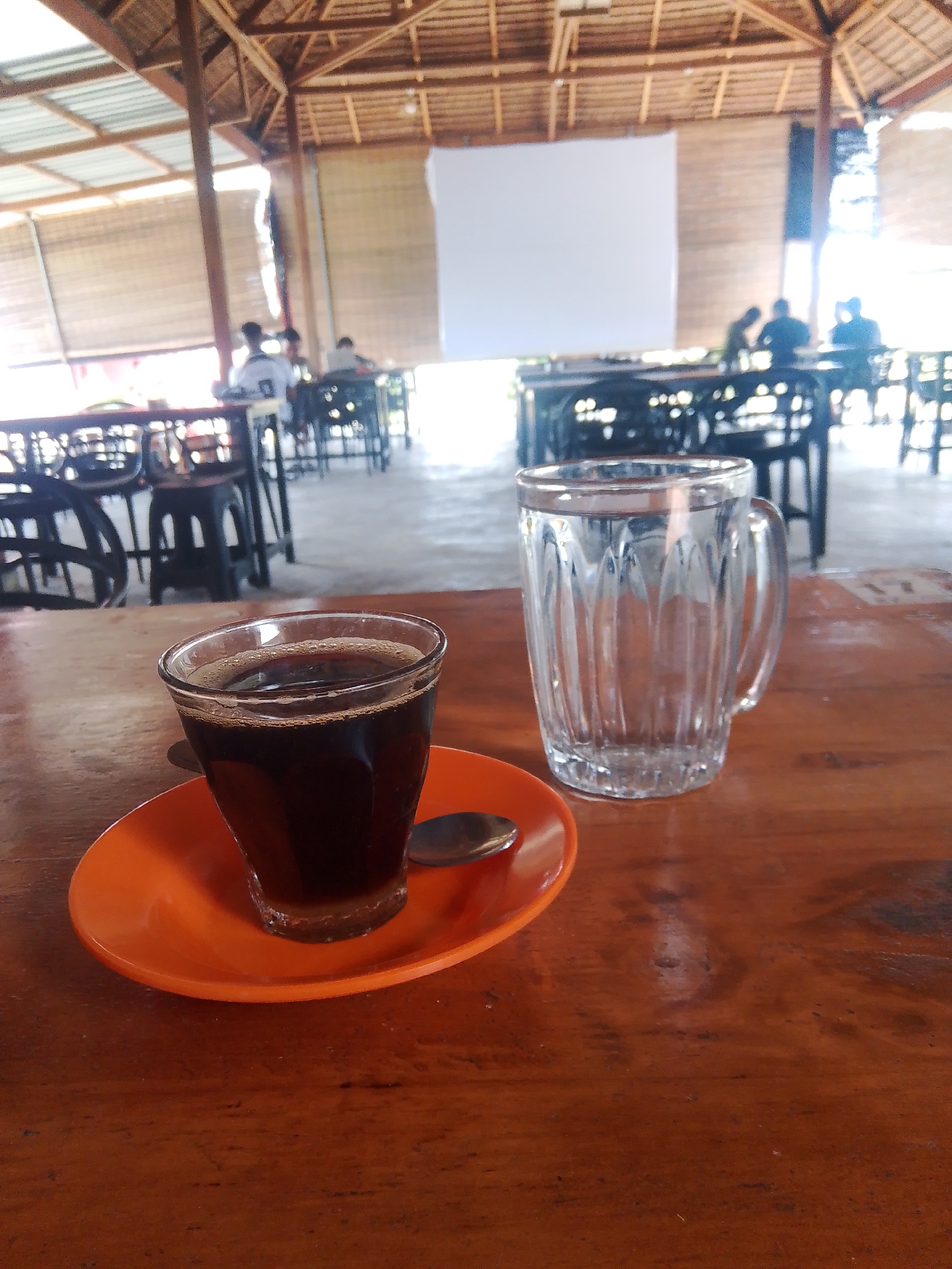 The image depicts a close-up view of a wooden table at an outdoor cafe. On the table, a dark liquid, likely coffee, sits in a clear glass on an orange saucer with a spoon. To the right, an empty glass mug with vertical ridges is visible. In the background, a roofed structure with bamboo blinds can be seen, suggesting a tropical or warm climate. Several tables and chairs are arranged under the roof, with blurred figures of people seated at them, indicating the cafe is in operation. The scene is set in Meunasahmee, Indonesia.