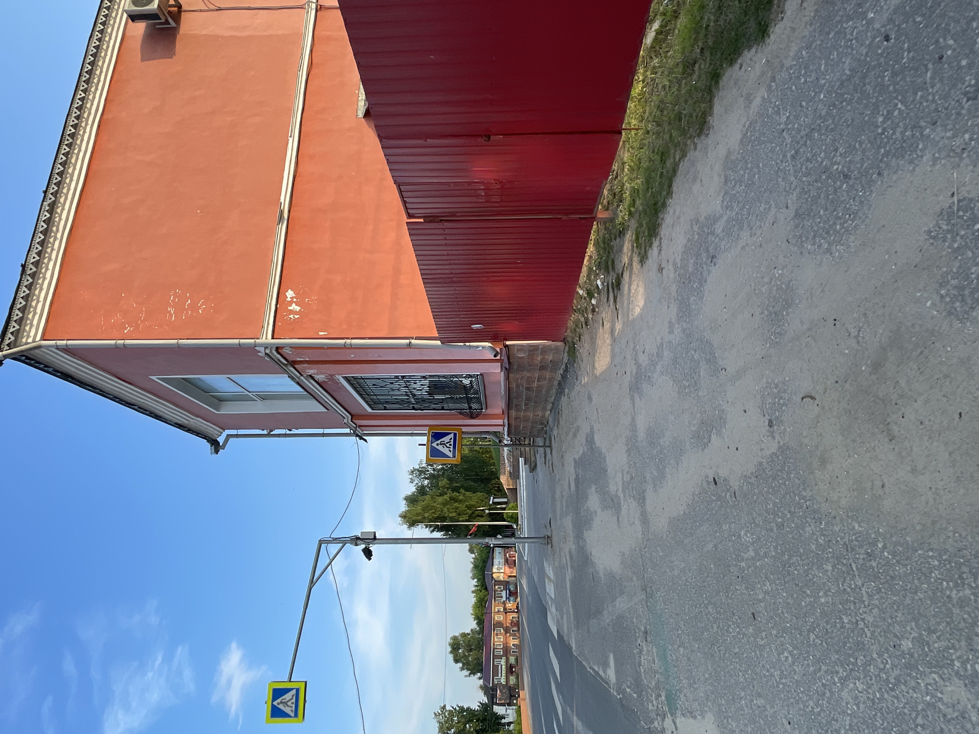 The image shows a street scene in the town of Krasnye Baki, Russia. In the foreground, a paved road with a pedestrian crossing is visible. To the right, a tall, bright orange building with white trim and decorative detailing on the roofline dominates the scene. A red metal fence runs along the side of the building. In the background, across the street, are several brick buildings with pitched roofs, suggesting a commercial or residential area. Trees are present behind these buildings and to the left of the orange building. Two pedestrian crossing signs are visible, one hanging from an overhead street lamp and another on a pole near the building. The sky is blue with a few wispy clouds, indicating daytime. An air conditioning unit is mounted on the side of the orange building. The overall impression is of a quiet street on a clear day.