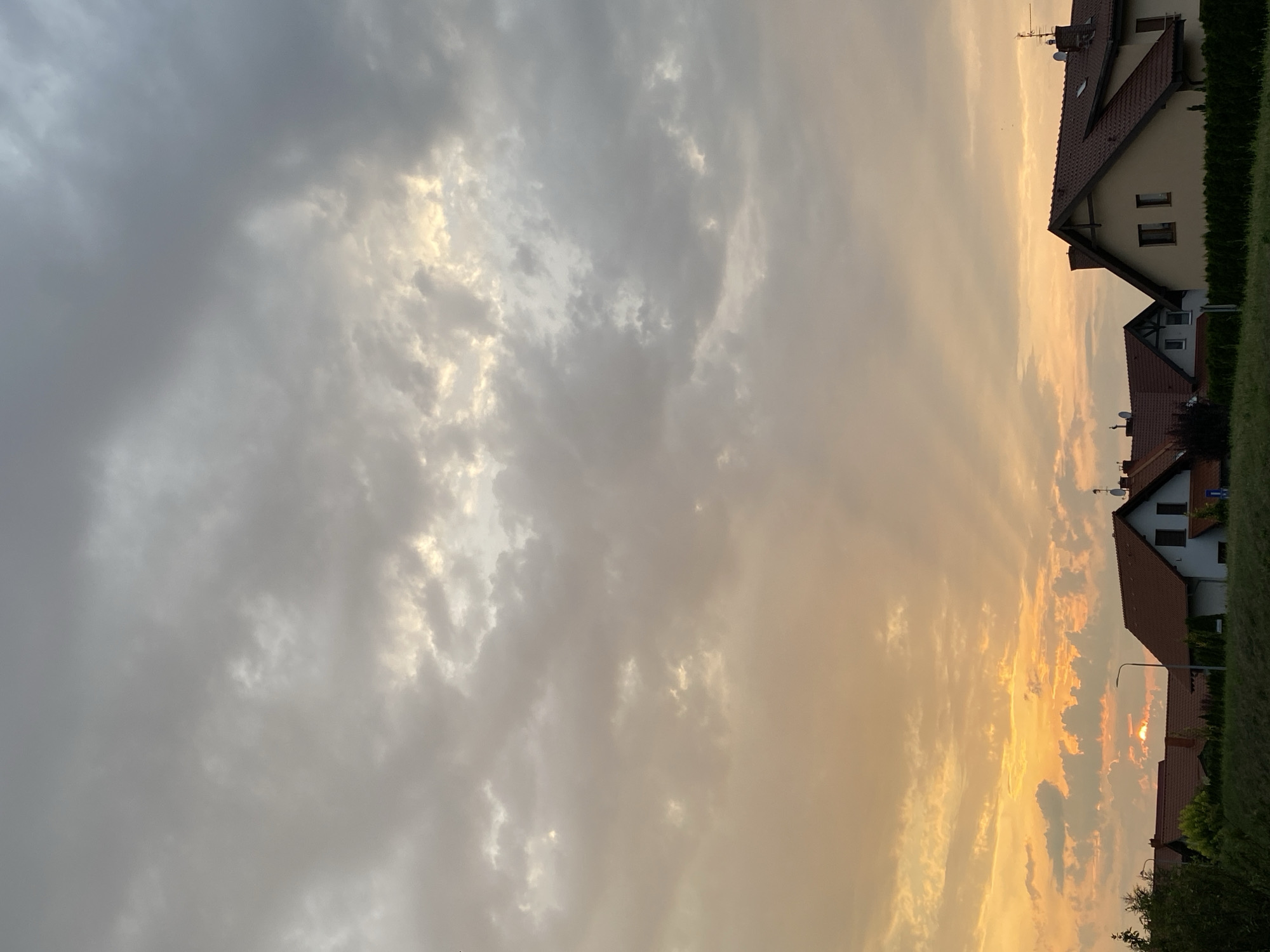 The image shows a suburban landscape in Buk, Poland, under a cloudy sky during what appears to be sunset. Several houses with red tile roofs are visible, lined up along a street or property boundary. The sky is filled with a mix of grey and orange clouds, with the warm hues of sunset filtering through. A street lamp is also present in the scene. The foreground consists of green grass and some foliage.