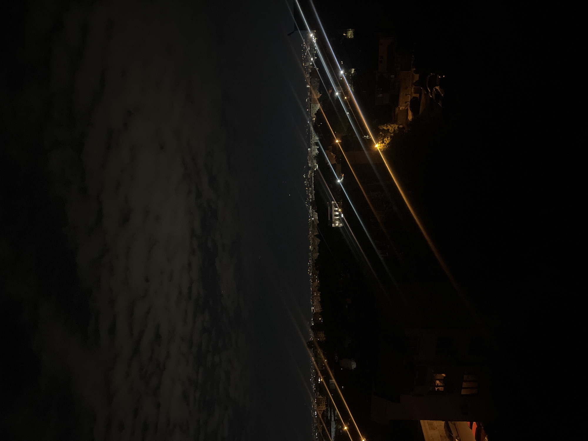 The image depicts a night view, likely taken from a high vantage point in Sofia, Bulgaria. The left side of the image shows a dark sky with scattered clouds. The center reveals a cityscape illuminated by streetlights and building lights stretching along a shoreline. The right side of the image features a dark foreground, partially obscuring some structures, with light streaks indicating possible traffic. The scene suggests an urban environment, with the city's lights contrasting against the night sky.