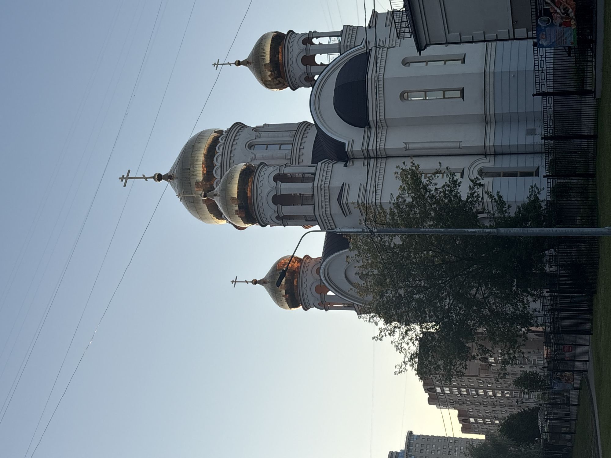 The image shows a large, white church with multiple golden domes, each topped with a cross. The church has a classic architectural style with arched windows and decorative elements. In the foreground, there is a tree with green foliage partially obscuring the view of the church. The background features a clear blue sky with a few power lines stretching across. To the right of the church, a black metal fence encloses an area, and beyond that, a modern apartment building with many windows is visible. The lighting suggests it is either early morning or late afternoon, indicated by the soft, warm light on the domes. There is no discernible activity or event taking place. The overall scene conveys a sense of serenity and urban architecture. No text is clearly visible.