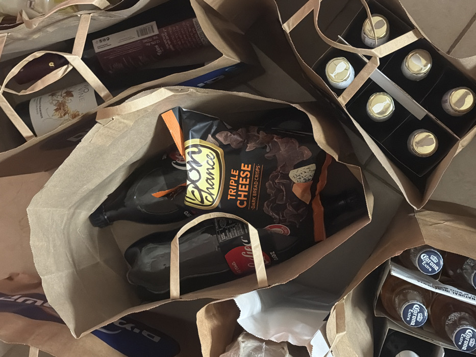 The image shows several brown paper shopping bags filled with various items. One bag contains a bottle of wine, a bottle of Coca-Cola Zero, and a bag of "Bon Chance Triple Cheese Dark Bread Crisps." Another bag holds a six-pack of small bottles with gold caps. A third bag contains multiple bottles of Corona Extra beer. Other items, including another bottle of wine, are visible in the background.