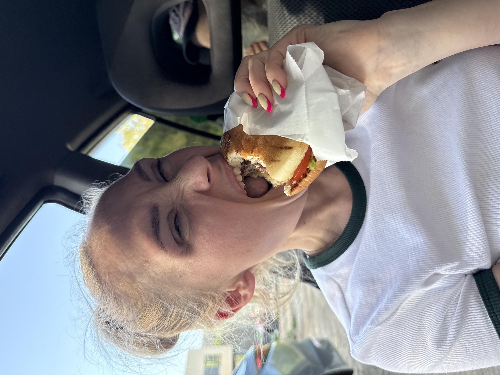 A woman is depicted eating a burger. She is inside a car, likely in the driver's seat, based on the position of the interior features of the vehicle. The woman is holding the burger in a white paper wrapper with her right hand. Her nails are manicured with a two-tone design. She's wearing a white t-shirt with a green trim. The location is presumed to be Chaikovsky, Russia. Further forensic details are not discernible.