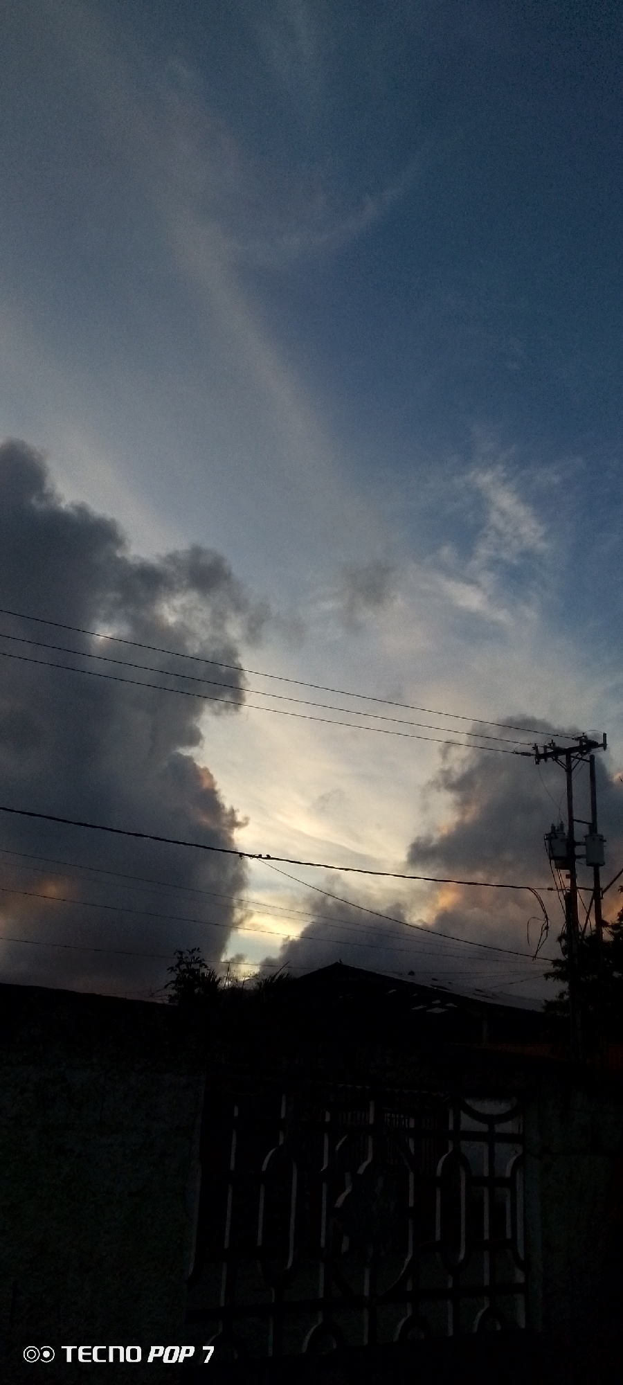 The image captures a dramatic sky scene, likely at sunset or sunrise, over Altagracia, Venezuela. The sky is dominated by large, dark, and heavy storm clouds on the left and right, creating a natural frame. Between these dark formations, lighter, wispy clouds are visible, illuminated by soft yellowish-orange light, suggesting the low angle of the sun. Patches of clear, deep blue sky can be seen in the upper regions.
Multiple power lines stretch horizontally across the mid-ground, with a utility pole and electrical equipment visible on the far right. Small dark specks, likely birds, are perched on some of the power lines. The bottom of the image is composed of dark, undifferentiated silhouettes of buildings, a patterned metal fence, and sparse vegetation, indicating an urban or semi-urban environment. The overall impression is one of impending or receding weather, with the sky showcasing a striking contrast between dark storm clouds and the softer light of dawn or dusk. A "TECNO POP 7" watermark is visible in the lower-left corner of the image.