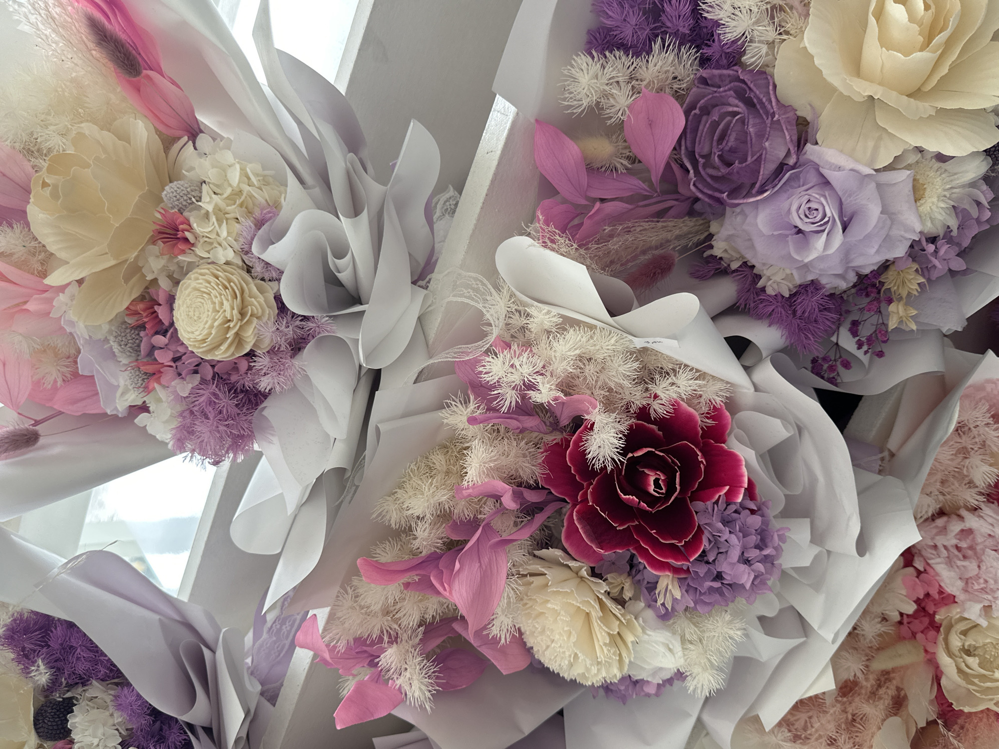 The image displays several dried flower bouquets arranged on white shelves. The bouquets feature a variety of dried flowers in shades of purple, pink, cream, and white, including roses, hydrangeas, and feathery accents. The wrapping paper is primarily white and pale purple. The background suggests an indoor setting, possibly a flower shop or a display area. There are no people visible in the image, and no specific landmarks or street views are present to indicate a precise location within Davao City, Philippines. The overall scene is one of still life, focusing on the artistic arrangement of dried floral elements.