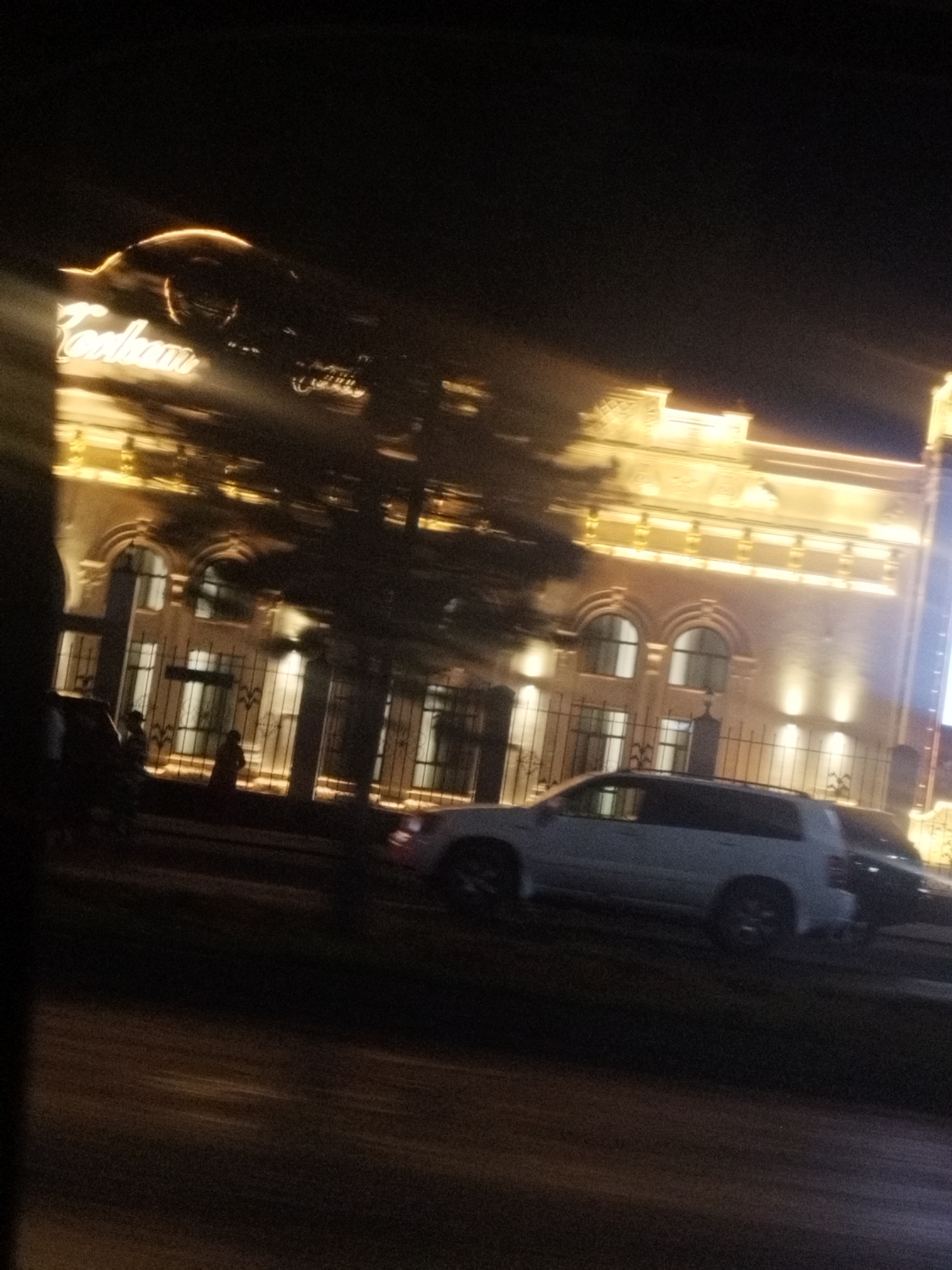 The image depicts a nighttime scene in Shymkent, Kazakhstan, showing a brightly lit building with ornate architecture. The building, illuminated by warm, yellow lights, features multiple arched windows and decorative elements along its facade. In front of the building, there is a black metal fence and several trees, their branches silhouetted against the light. A white SUV is parked in the foreground, and another dark vehicle is partially visible behind it. Two figures, one appearing to be wearing a dark coat and the other a darker outfit, are standing near the fence. The street in front of the building is visible and appears to be paved. The overall atmosphere is one of a well-lit urban environment at night.