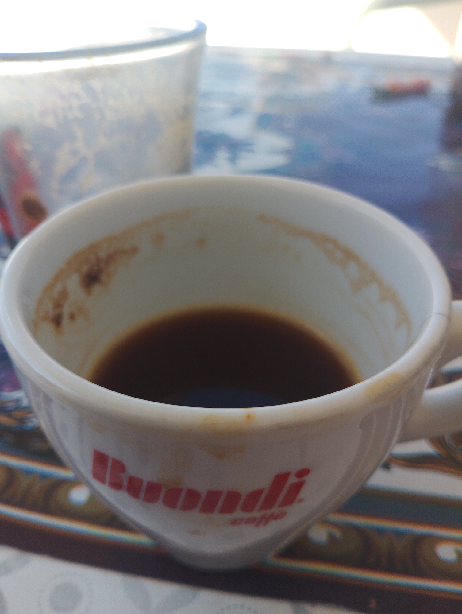 A close-up shot shows a white ceramic coffee cup with a small amount of dark liquid remaining at the bottom. The inside rim of the cup has some dried residue. The exterior of the cup features red lettering that appears to spell "Buondi" with "caffè" below it. The cup is positioned on a patterned surface, possibly a tablecloth, with a blurred glass in the background. The scene is brightly lit, suggesting it might be daytime. The location is identified as Lakki, Greece, though no specific landmarks or contextual details within Lakki are visible in the image.