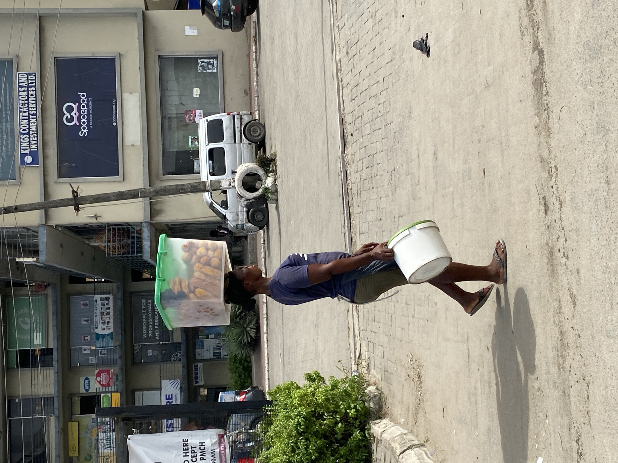 A man in Lagos, Nigeria, walks on a sidewalk carrying a container on his head filled with long, tan food items. He also carries a white bucket. The man wears a blue shirt, green shorts, and dark flip-flops. He is walking past a multi-story building with various storefronts and signage, including advertisements for "Spacepod" and "KINGS CONTRACTORS AND INVESTMENT SERVICES LTD." A white van is parked nearby, next to a stack of tires.