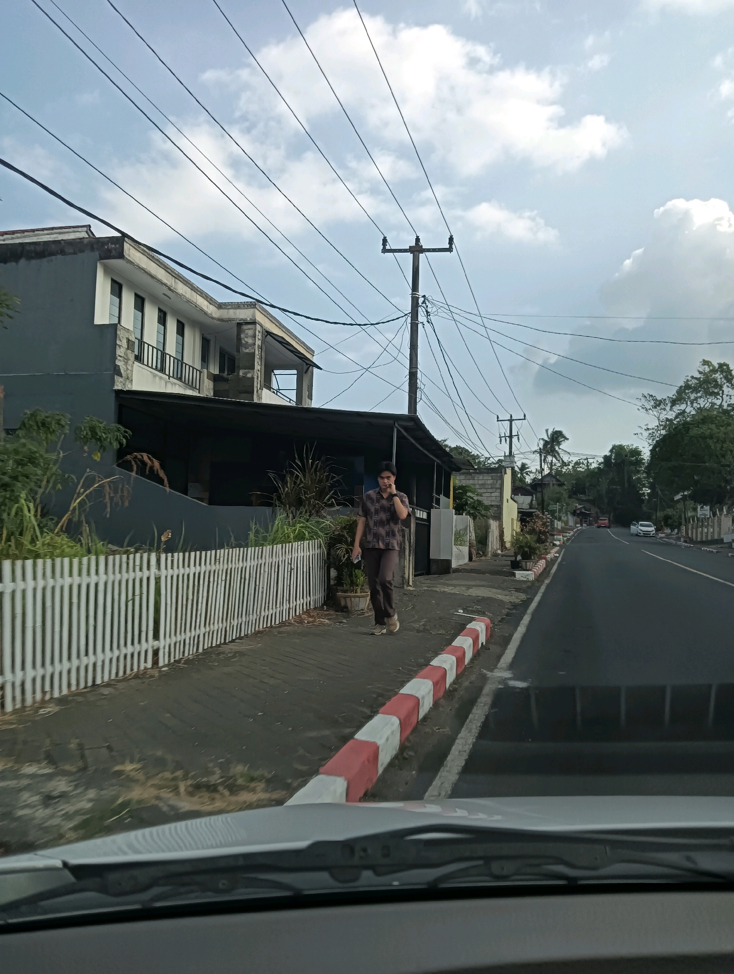 The image shows a street scene in Tomohon, Indonesia, with a person walking on the sidewalk next to a road. The person, who appears to be male, is wearing a patterned short-sleeved shirt and dark trousers. He is walking towards the right side of the frame and has his right hand near his ear, suggesting he might be on the phone.
To the left of the sidewalk is a white picket fence, behind which is a two-story building with dark gray and stone facade elements. The building has multiple windows and balconies. Beyond the building, there are various plants and trees.
The road is paved and curves to the right in the distance. There are red and white striped concrete barriers along the edge of the sidewalk and road. In the distance, other vehicles are visible on the road, including a white car and an orange car. Overhead, numerous power lines crisscross the sky, supported by utility poles. The sky is partly cloudy. The foreground shows the dashboard and windshield wipers of a vehicle, indicating the photo was taken from inside a car.