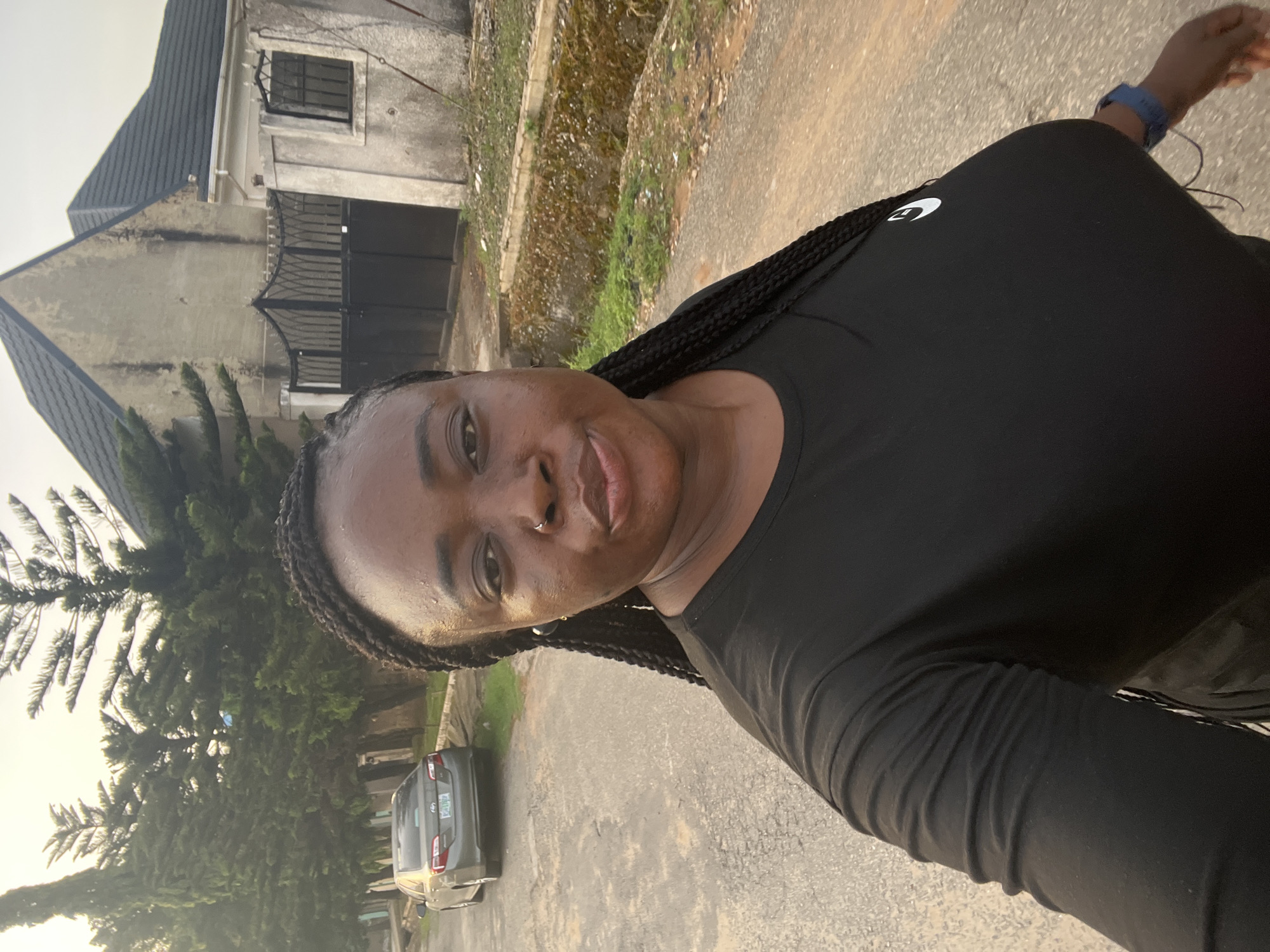 A person with braids is taking a selfie on a paved street in a residential area. The person is wearing a black long-sleeved shirt with a white logo and a blue wristwatch. Behind the person is a house with a black gate and a grey car parked on the street, beside trees. The city of Abuja, Nigeria, is not explicitly evident in the image but may be inferred from the context of the provided information.