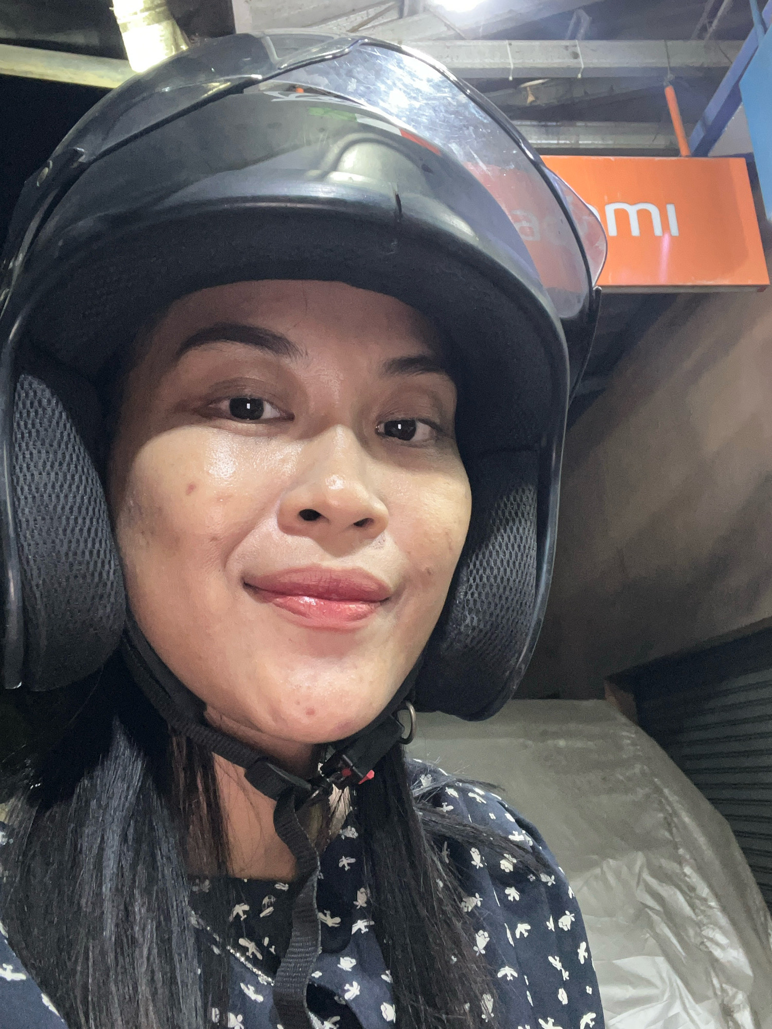 A medium shot photo shows a woman wearing a black helmet and a dark blue shirt with white floral patterns. She is looking at the camera and smiling. Her hair is long and black, and it falls over her shoulders. She has brown eyes and her lips are painted with a pinkish shade of lipstick. The background is blurry and appears to be an outdoor setting at night. There is a sign in the background with the word "mi" visible in white letters on an orange background. The overall impression is a casual selfie taken in a public place.