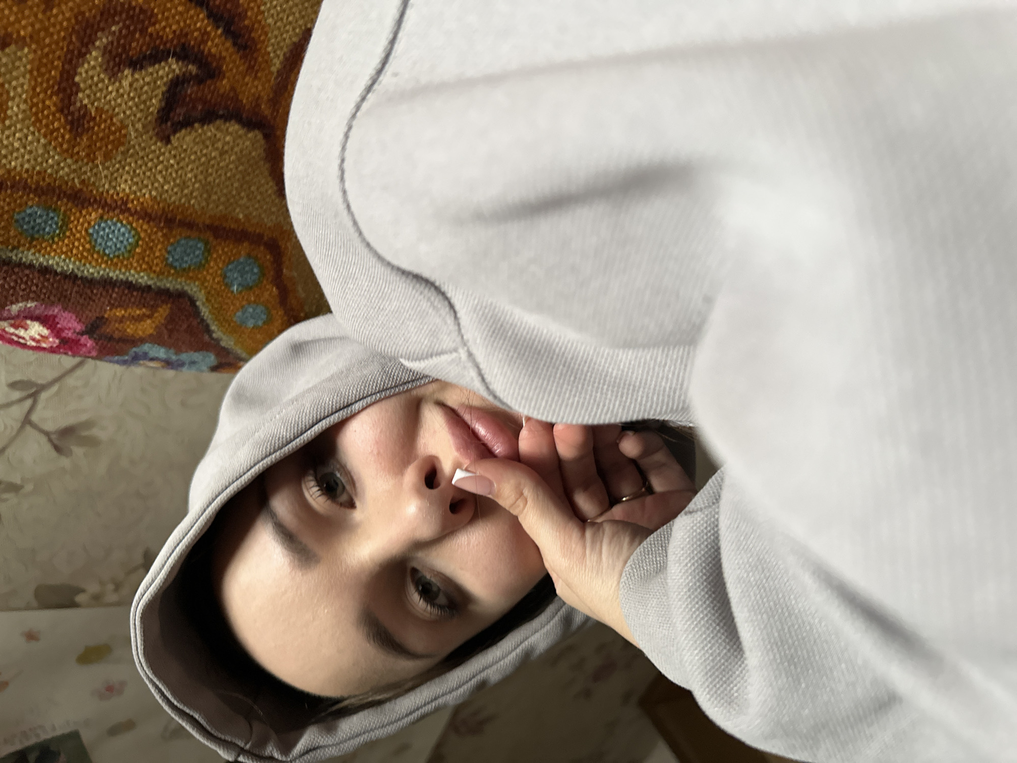 A woman with dark eyebrows and light eyes is wearing a light gray hoodie. The camera angle is from above, looking down at her face. Her right hand is raised, with her fingers near her lips. Part of a patterned couch or chair is visible in the background. The location cannot be determined from the image.