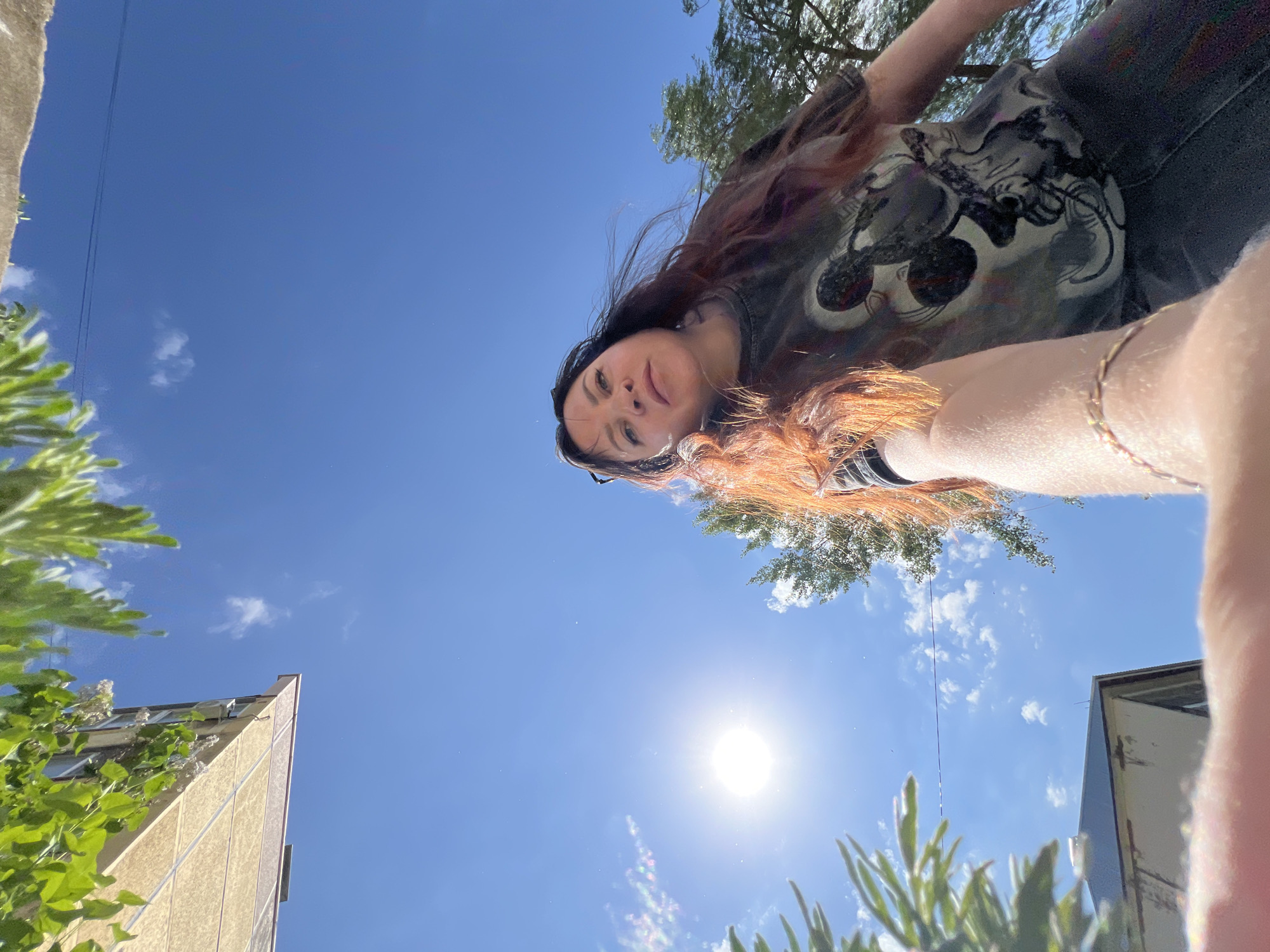 A low-angle, outdoor photograph depicts a woman with long, wavy, auburn-tinged brown hair looking downward at the camera. She wears a black T-shirt with a graphic design. A delicate gold bracelet is visible on her wrist. The background is a bright blue sky with a few scattered clouds and the sun providing bright lighting. Parts of buildings with foliage are visible in the lower left and right corners of the frame. The location is Noviy, Russia.