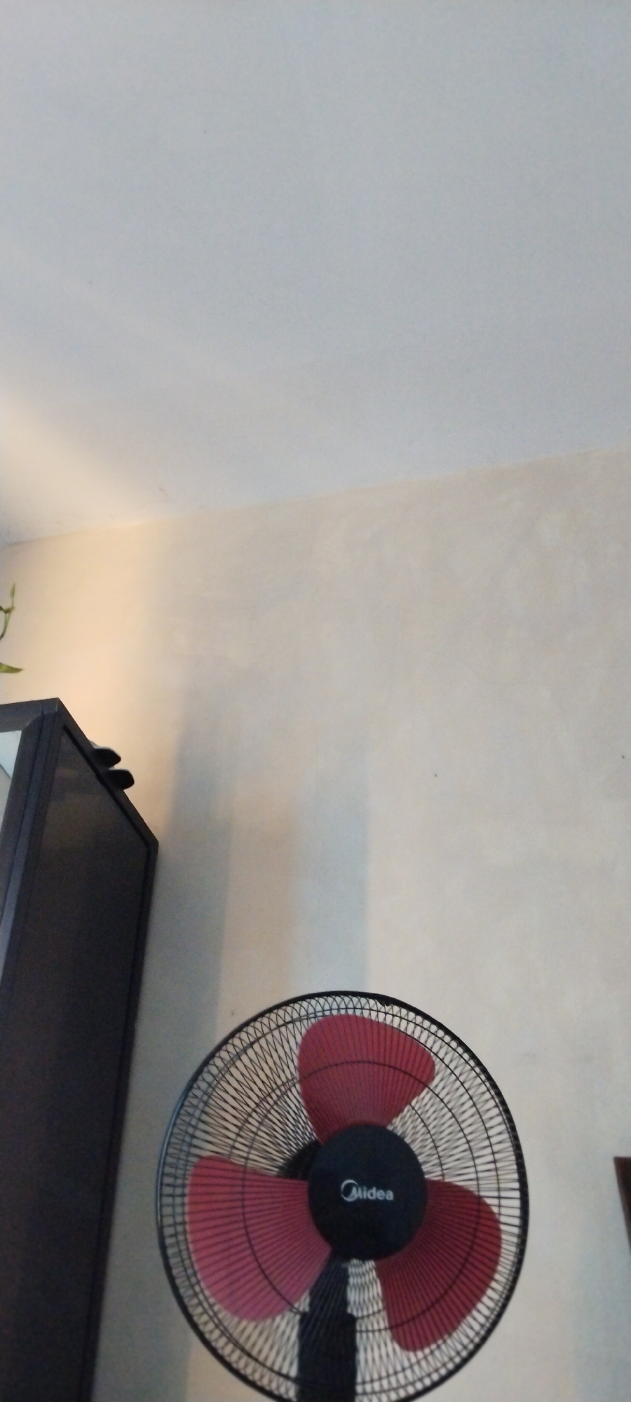 The image shows a close-up of a black oscillating fan with red blades, positioned in front of a neutral-colored wall. The fan's motor housing is black with a white "Midea" logo. The fan is the primary subject, occupying the lower right portion of the frame. To the left, a dark-colored cabinet or piece of furniture is partially visible. The background is a wall that appears to be lit from the left, casting a subtle shadow. The top of the image shows a ceiling. There is no discernible activity taking place, and the scene is static. The lighting suggests it could be daytime indoors. No specific location cues beyond the general indoor setting are evident, and no emotions are conveyed. The visible text is the brand name "Midea" on the fan.