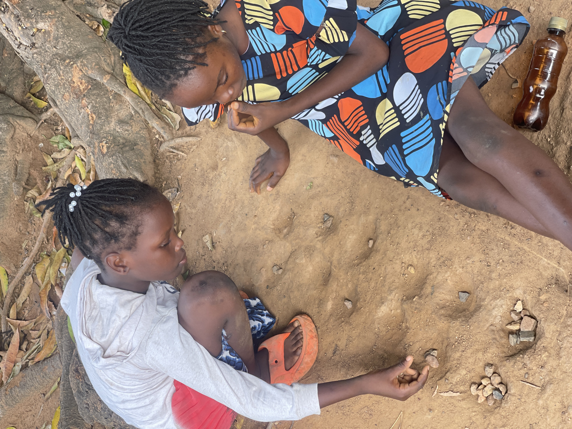 Two young Black girls sit on a reddish-brown dirt ground outside under trees. One is lying on her side wearing a colorful short-sleeved dress in a pattern of blue, yellow, orange, and white geometric shapes, next to a brown plastic bottle. The other girl is sitting up with her knees bent, wearing a white hoodie and a red and blue patterned garment. She has on one orange sandal and is playing with small stones. The location is Akwanga, Nigeria.