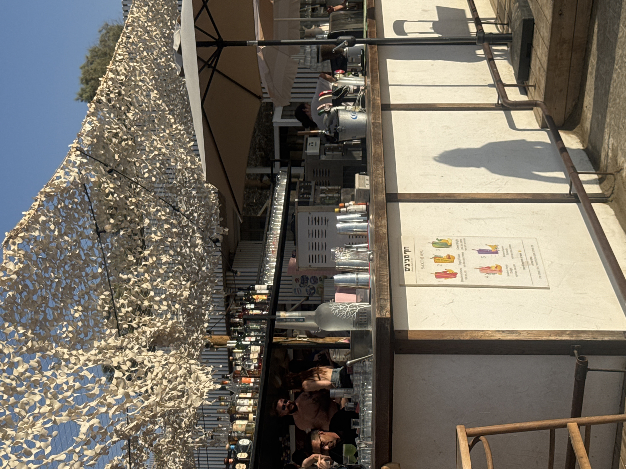The image depicts an outdoor bar located at Metzitzim Beach (חוף מציצים) in Tel Aviv, Israel, during daytime under clear blue skies, with sunlight casting strong shadows. A light-colored camouflage netting, functioning as a sunshade, spans overhead, partially obscuring the sky.
The central feature is a bar counter with a wooden top and a white paneled base. On the counter, various bar essentials are visible, including numerous empty glasses, metal shakers, and a prominent clear bottle with a tree design (likely a spirits bottle). Behind the bar, shelves are densely stocked with an assortment of liquor bottles.
Several individuals are present:
* Behind the bar, a man with a beard and sunglasses, wearing a light shirt, interacts with someone.
* Another woman with long dark hair, wearing a dark top and light shorts, is also behind the bar.
* To the left, a man wearing glasses and a dark shirt is seated at the bar, with a green beverage in a glass before him.
* Further back on the right, a person with dark hair tied back is working amidst various stainless steel equipment and a large beige patio umbrella, which provides additional shade.
A white sign displaying "חוף מציצים" (Metzitzim Beach) in Hebrew and a "SMOOTHIE MENU" is affixed to the front of the bar counter. The menu features colorful illustrations of six different smoothie options, including "Strawberry Banana," "Tropical Fusion," and "Green Delight." In the foreground, a wooden bar stool is visible on the far left. The ground is a mix of wooden decking and a light-colored surface, with a dark metal railing running along the front. Elongated shadows of people and a chair are cast onto the ground and the light-colored wall, indicating the sun's position.